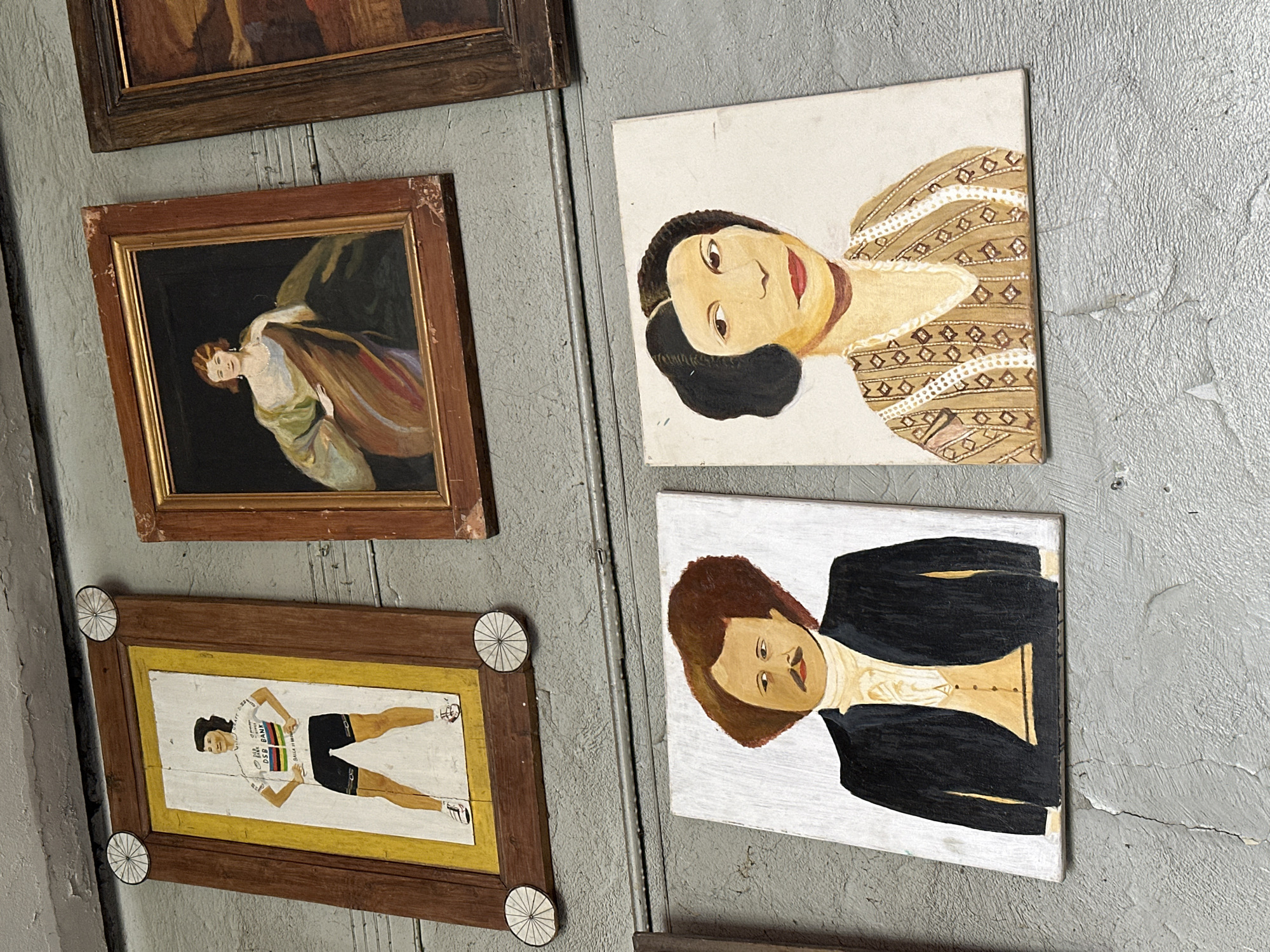 The image shows a wall with several framed paintings hanging on it. The wall is gray and appears to be textured. From top to bottom, paintings are hung in vertical rows, positioned to the left and to the right.
On the left, from top to bottom, there's:
- A painting in a dark brown frame.
- A painting of a woman with light skin and dark hair, draped in fabric, in a brown wooden frame.
- A painting of a female cyclist in a white, yellow, black, and red racing uniform in a brown wooden frame. It has white wheel-like decorations on each corner.
On the right, from top to bottom, there's:
- A portrait painting of a dark-haired woman in a brown and white dress.
- A portrait painting of a brown-haired person in a black suit and white shirt.
- The lower part of a framed painting.
This scene could be set in an art gallery or a private collection. Given the context and the prompt's indication of Tbilisi, Georgia, it's plausible this is a location there.