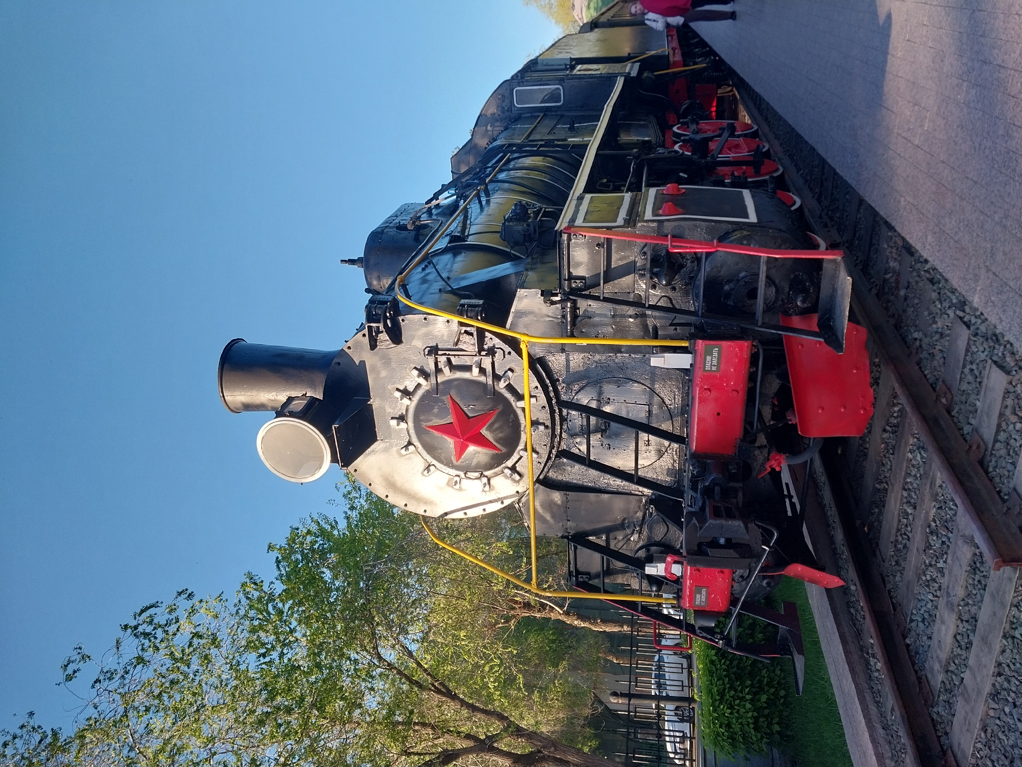 The image shows a Soviet-era steam locomotive in Orenburg, Russia. The locomotive is black with red accents, notably a large red star on the front. Yellow safety barriers are visible. The locomotive is on display on a section of railway track. Behind the locomotive are trees, a fence, and a building. The sky is clear and blue. In the upper right corner, part of a person is visible on a paved area.