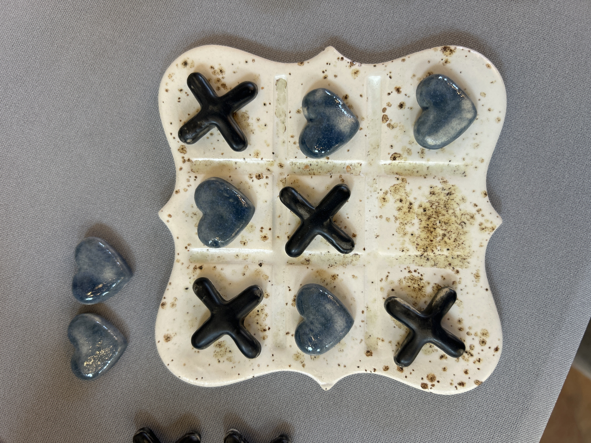 The image shows a tic-tac-toe game with a ceramic board and heart-shaped and 'X' shaped playing pieces. The board is off-white with brown speckles, and it holds five black 'X' pieces and three blue heart pieces. Two additional blue heart pieces are next to the board. The game appears to be in progress or recently finished, with an empty square in the middle row on the right side. The game and extra pieces are on a gray fabric surface. The location in the city of Sofia, Bulgaria is unknown.