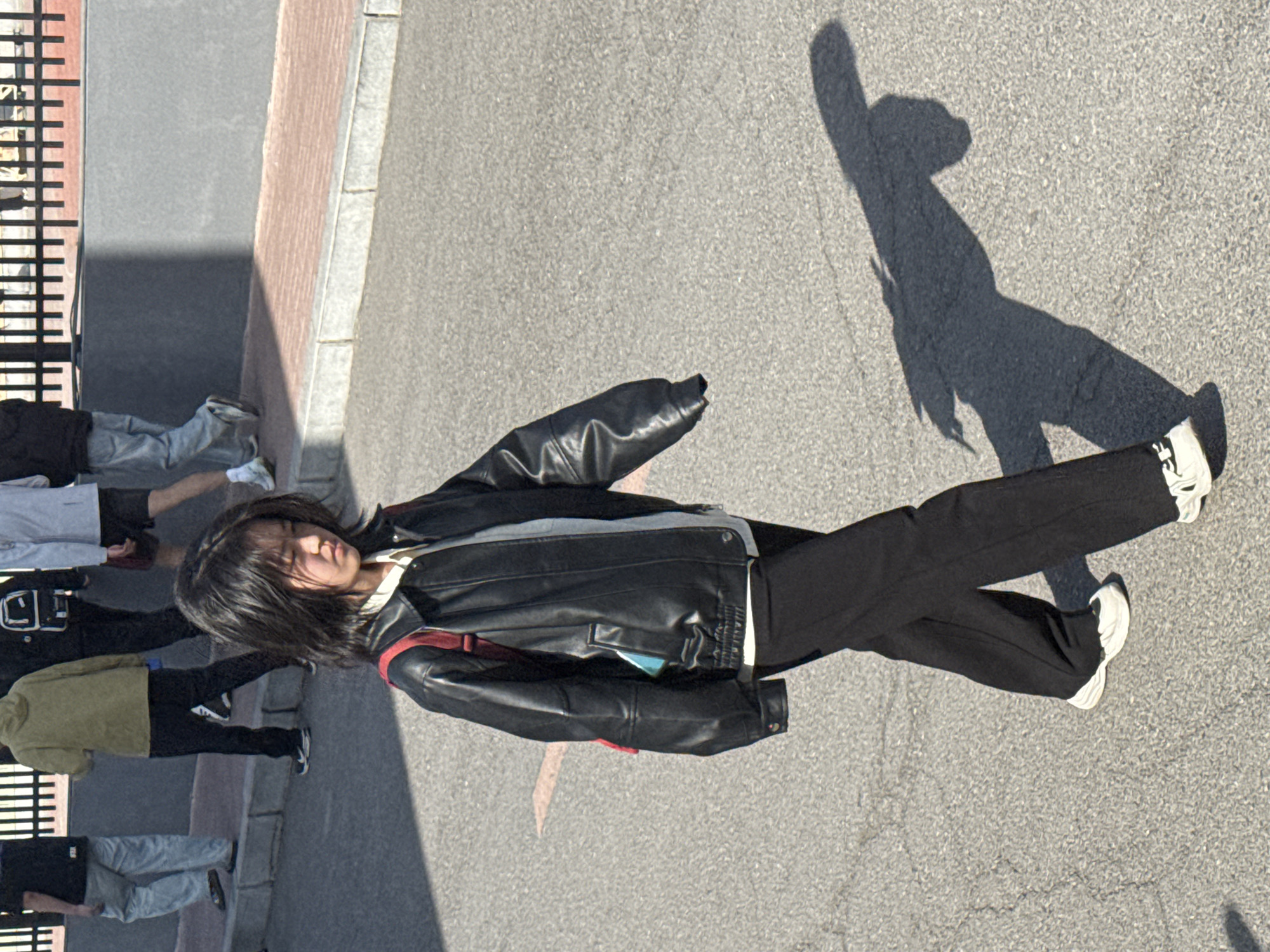 This image, taken in Zhongtun, China, captures a scene on a sunny day in what appears to be an urban or suburban street or pedestrian area. The main subject is a young person, likely a teenager or young adult, with dark, medium-length hair, facing slightly upwards with their eyes closed or squinting, possibly due to bright sunlight. They are wearing a black leather jacket over a light-colored top, dark pants, and white sneakers. A red strap, likely from a bag, is visible across their shoulder.
The person is captured in mid-stride, with their body angled slightly. To their right, a long, distinct shadow of the individual is cast on the light gray asphalt, indicating the sun is low in the sky, suggesting either early morning or late afternoon. The ground is composed of a textured gray asphalt, adjacent to a reddish-brown paved strip, and a gray curb. On the far left of the frame, a black metal fence or railing runs parallel to the ground.
In the background on the left, several other people are visible, mostly their legs and lower torsos, also appearing to be casually dressed and either standing or walking. The overall impression is a bustling public space on a clear, bright day. No discernible text is visible on any objects or clothing.