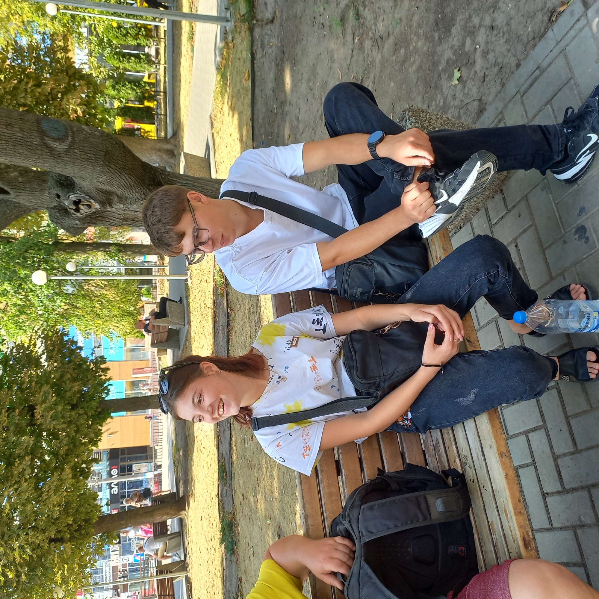 Two young people, a girl and a boy, are sitting on a wooden bench in a park. The girl, on the left, is wearing a white t-shirt with colorful graphic prints and dark jeans. She has sunglasses on her head and a crossbody bag over her shoulder. She is smiling and looking to her left. The boy, on the right, is wearing a white t-shirt and dark jeans. He has glasses and a crossbody bag. He is looking to his right. A black backpack is on the bench between the girl and another person whose legs are partially visible on the far left. A blue water bottle is on the ground in front of the girl. The park appears to be in a city, with trees and a paved walkway in the background. The scene is illuminated by natural sunlight. The location is the City Garden in Kropyvnytskyi, Ukraine.