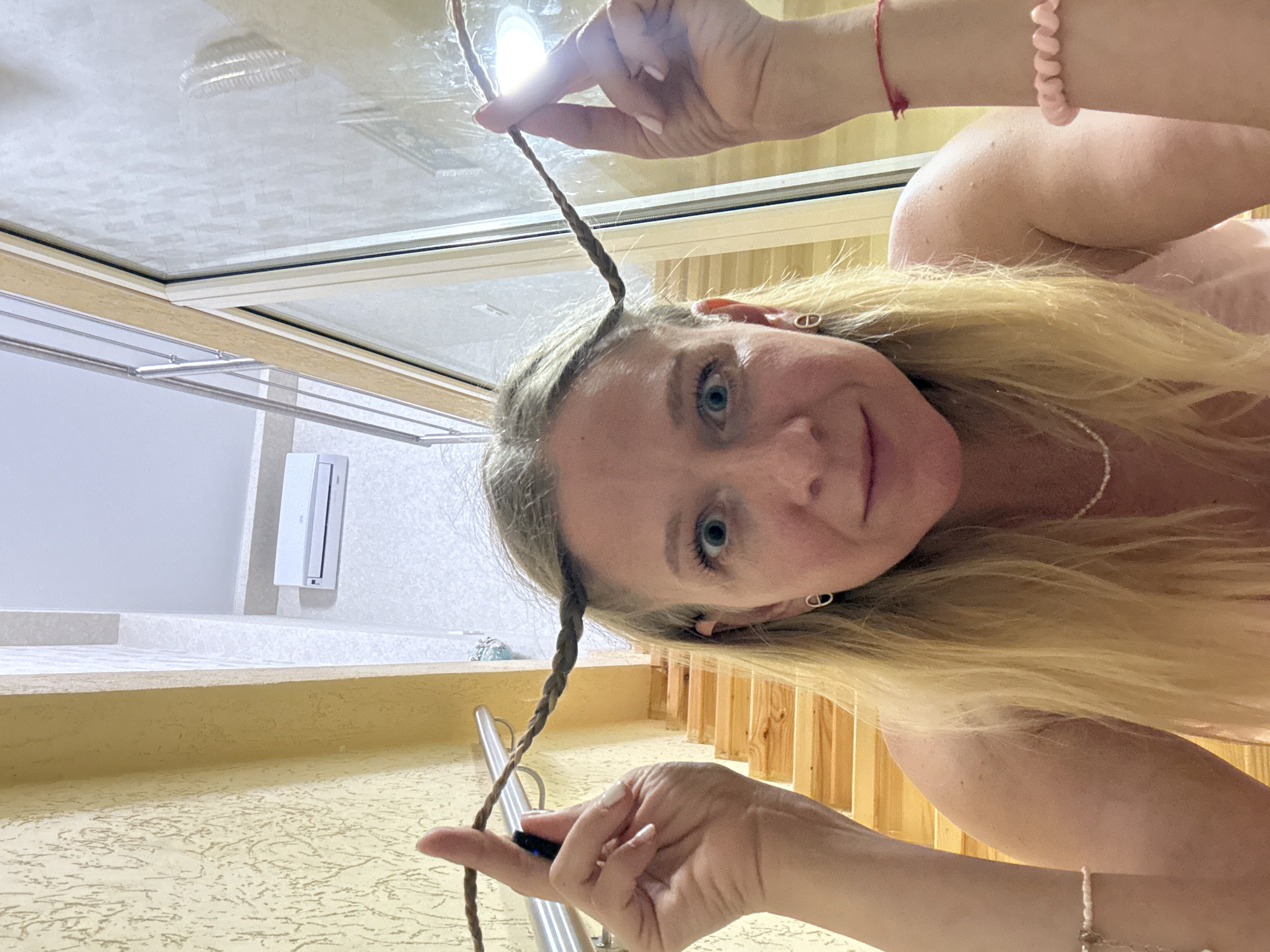 A woman with blonde hair and blue eyes is pictured from the chest up. She is looking directly at the camera with her hair twisted into two braids that extend outwards from her forehead. The woman is wearing a pink top and pearl necklace. She has earrings and bracelets on her arms. In the background, there is a wooden staircase and a large window. The lighting suggests an indoor setting. The overall scene appears to be a casual, indoor environment.