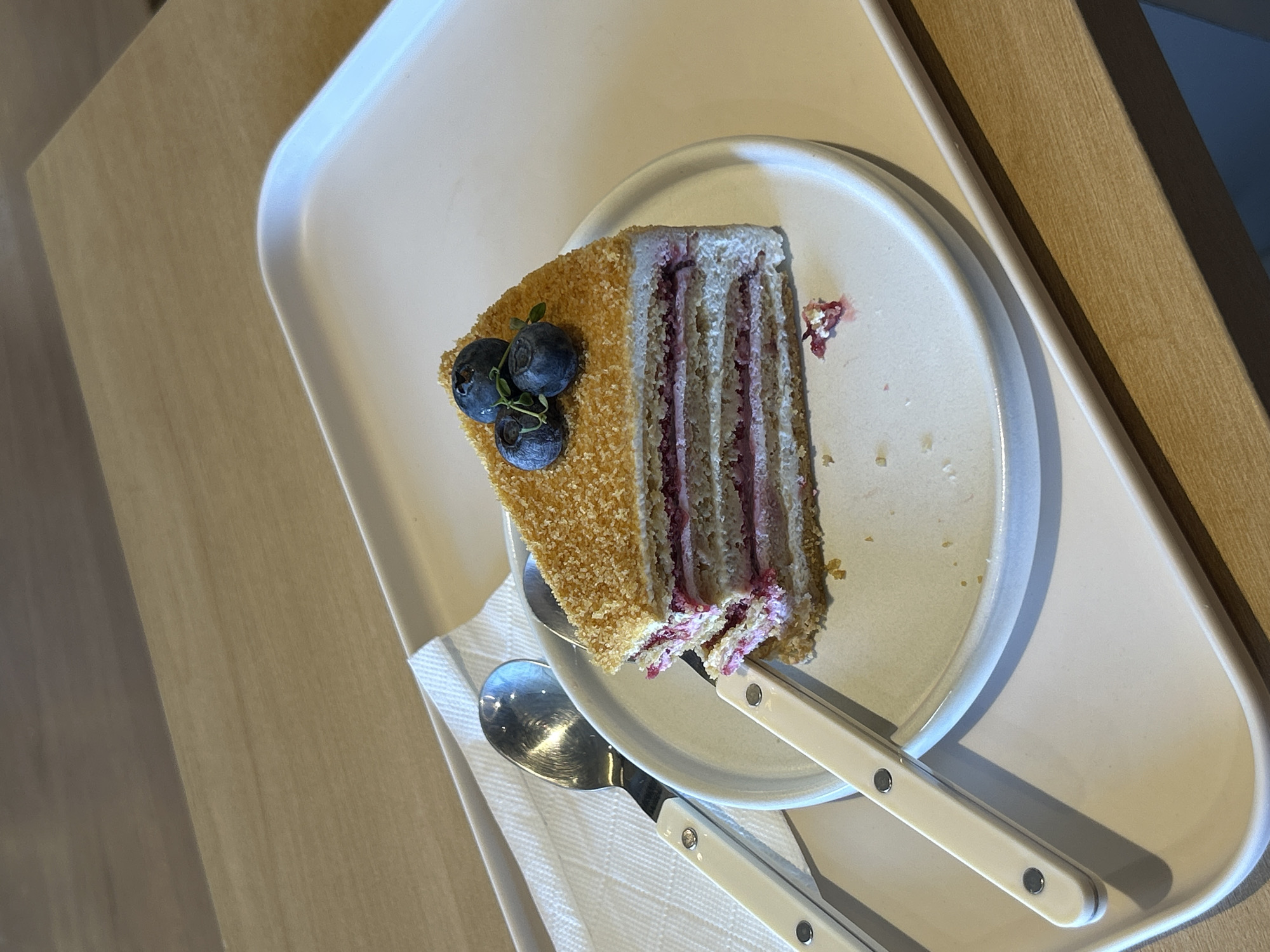 A slice of layered cake with a berry filling and a crumb coating is presented on a white plate. Three blueberries and a sprig of greenery garnish the top of the cake. A spoon and a fork, with white handles, are placed beside the plate on a white napkin. The plate rests on a larger, white rectangular tray, which is positioned on a light-colored wooden surface. This scene is located in the city of Chiang Mai City Municipality, Thailand.