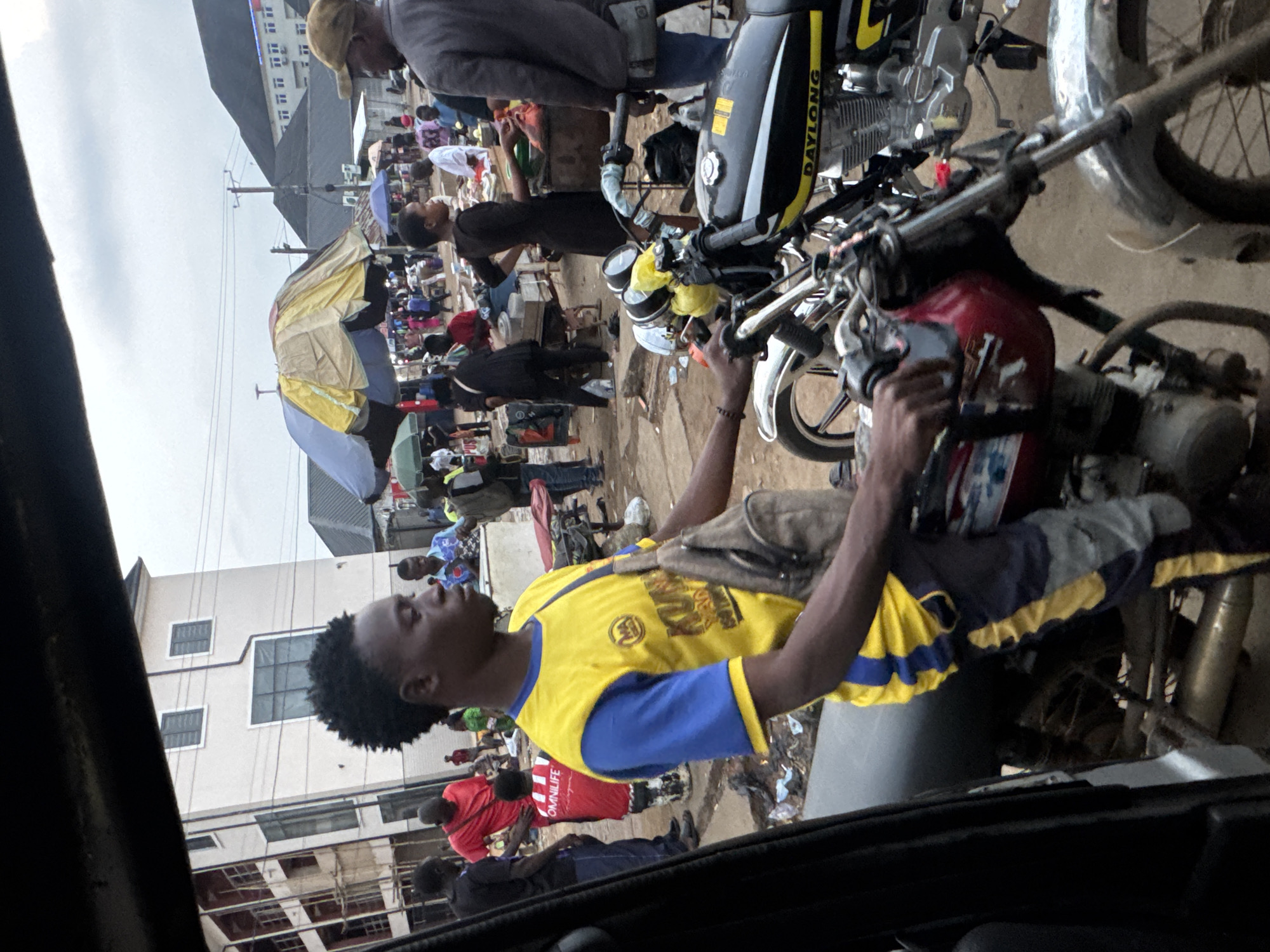 The image depicts a bustling street market scene in Obinze, Nigeria. In the foreground, a young man wearing a yellow and blue t-shirt and dark pants is seated on a red and black motorcycle. He has a backpack slung over his shoulder and appears to be looking to his right. Another motorcycle, primarily black with yellow accents and the brand name "DAYLONG" visible, is parked alongside his.
The background reveals a crowded market with numerous people engaged in various activities. Stalls with colorful umbrellas are set up, suggesting the sale of goods. Buildings, including a multi-story structure with balconies under construction and a white building with multiple windows, are visible in the distance. The ground is unpaved and appears dusty, with some debris scattered around. The sky is overcast, indicating a cloudy day. The overall impression is one of a vibrant and active community market.