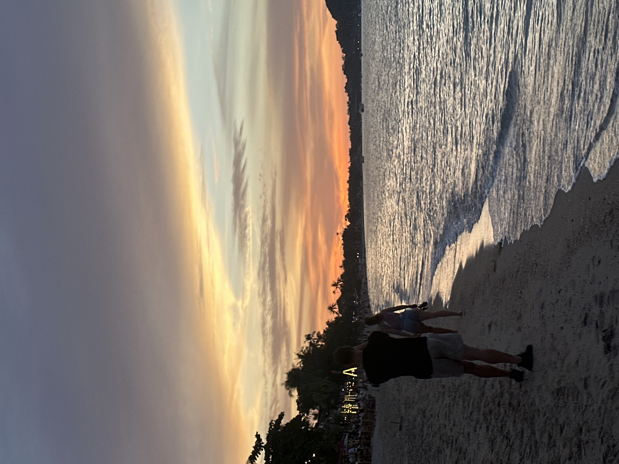 This is a full shot, eye-level, vertical photograph of a beach in Ko Samui District, Thailand, during sunset. The sky is filled with streaks of orange, yellow, and purple clouds. The ocean on the right side of the image is calm with gentle waves lapping at the shore. On the left side of the image, there is a sandy beach lined with palm trees and some buildings with lights. Two people are walking on the beach towards the water. The person in the foreground, facing away from the camera, is a male wearing a black t-shirt and gray shorts. The person behind him is a female wearing a light-colored tank top and denim shorts. The lighting suggests it is twilight.