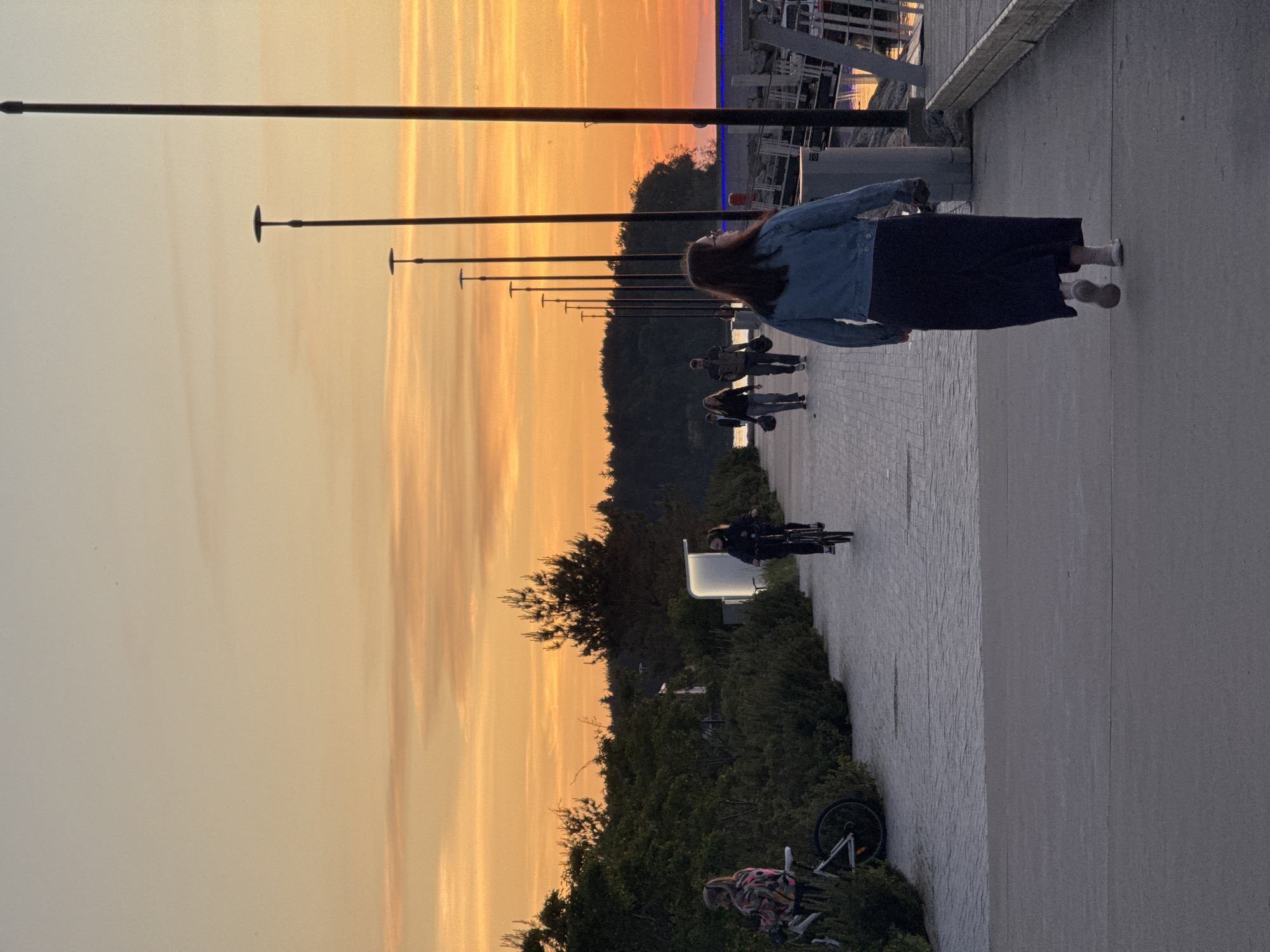 This photograph captures a sunset scene along the Tallinn Sea Promenade in Tallinn, Estonia. The sky is painted with vibrant hues of orange and yellow, indicative of the evening twilight. In the foreground, a woman with long dark hair, wearing a blue denim jacket and a dark skirt, is walking away from the camera on a paved walkway. Behind her, several other individuals are strolling or cycling. To the left, two people are seen on bicycles near a line of dense green bushes. Further along the promenade, a row of tall, slender lampposts stretches into the distance, silhouetted against the colorful sky. The walkway itself is paved with light-colored stones, transitioning to a darker concrete surface in the immediate foreground. On the right side of the image, a wooden pier with railings is visible, with structures and reflections of light in the water. The background features a line of trees and a body of water. The overall atmosphere is serene and captures a moment of leisure at the waterfront.