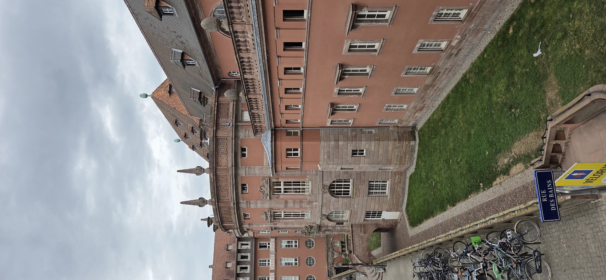 The image captures an exterior view of a building complex in Strasbourg, France, specifically the Palais de Justice. The architecture features salmon-pink facades, sandstone accents, and a variety of windows, some arched and some rectangular. Decorative elements like finials and ornate cornices are visible on the building's upper levels. A section of the building has a curved facade with decorative stonework above a row of arched windows. Adjacent to this is a more angular section with a pitched roof and dormer windows.
In the foreground, a paved area is lined with numerous bicycles parked in racks. A blue street sign reading "RUE DES BAINS" is prominently displayed, indicating the street name. Below this sign, a yellow sign with a cyclist icon and the word "VÉLODROME" is partially visible. A grassy area slopes down from the building towards the paved area with the bicycles. The sky is overcast with grey clouds. The scene suggests a public space, possibly a university campus or a municipal building, given the presence of bicycles and directional signage.