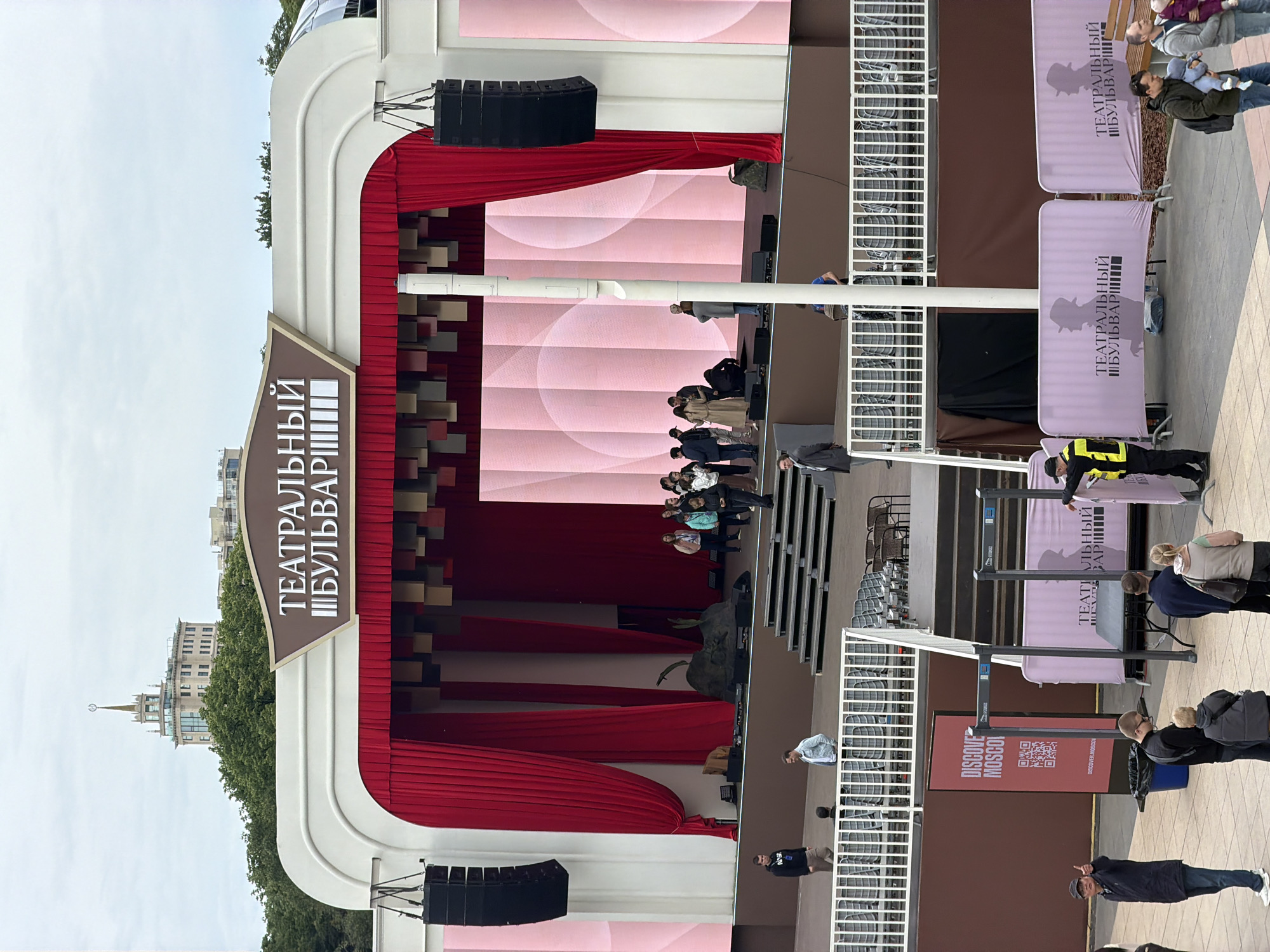 Here is a detailed description of the image:
**Scene:** The image features an outdoor stage, likely set up for an event or performance. The stage backdrop is pink with a patterned design. Red curtains are draped around the stage structure, which is primarily white and brown. Signage indicates that the event is taking place on the "Theater Boulevard," likely in Moscow, Russia.
**People:** Several individuals are visible. Some are standing on the stage, possibly performers or participants. Others are seated in rows of chairs set up in front of the stage, suggesting an audience. Some people are standing or walking in the foreground.
**Objects:** Besides the stage and seating, there are sound systems (speakers) mounted on either side of the stage. Banners with similar signage to the stage are also visible.
**Actions/Interactions:** People on stage are grouped together and may be preparing for or concluding a performance. Audience members are observing the activity on stage.
**Environment:** The backdrop includes a glimpse of buildings, indicating an urban setting, likely Moscow. The sky is overcast, suggesting a cloudy day. The location is on the Theatrical Boulevard of Moscow, Russia.