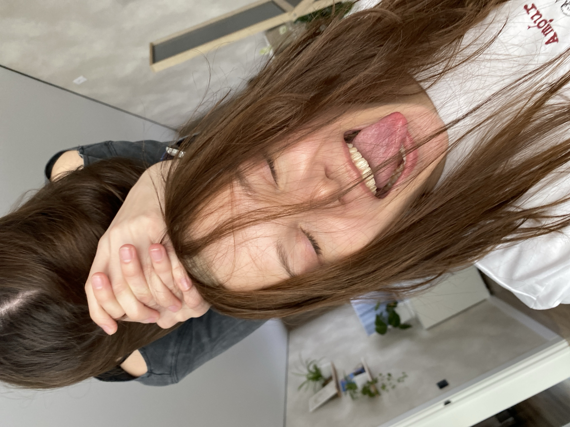 The image depicts a young woman indoors, lying on her back, possibly on a bed. She has long brown hair that partially covers her face. Her eyes are closed, and her mouth is open with her tongue sticking out. She is wearing a white t-shirt with some writing on it, partially obscured by her hair. Another person, with long brown hair and wearing a gray top, has their hand near the woman's face. The room appears to be a bedroom or living space, with a white door and a glimpse of plants on the floor near the wall. The location is unspecified, there is no mention of Votkinsk, Russia.