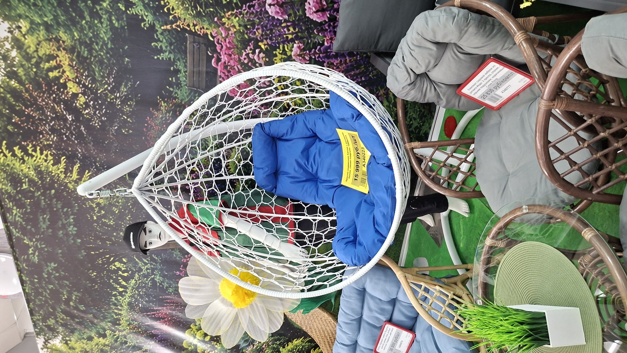 This photo, taken in Chaikovsky, Russia, showcases a collection of furniture pieces. A white hanging chair with a net-like design holds a blue cushion. Inside the chair, a mannequin wears a black hat, white top, and black pants. Next to it, brown rattan chairs feature grey cushions and red price tags. A glass-topped table holds artificial green grass in a white pot. The backdrop is a garden scene with green foliage and pink flowers. Price tags indicate the costs of the items in Russian rubles.