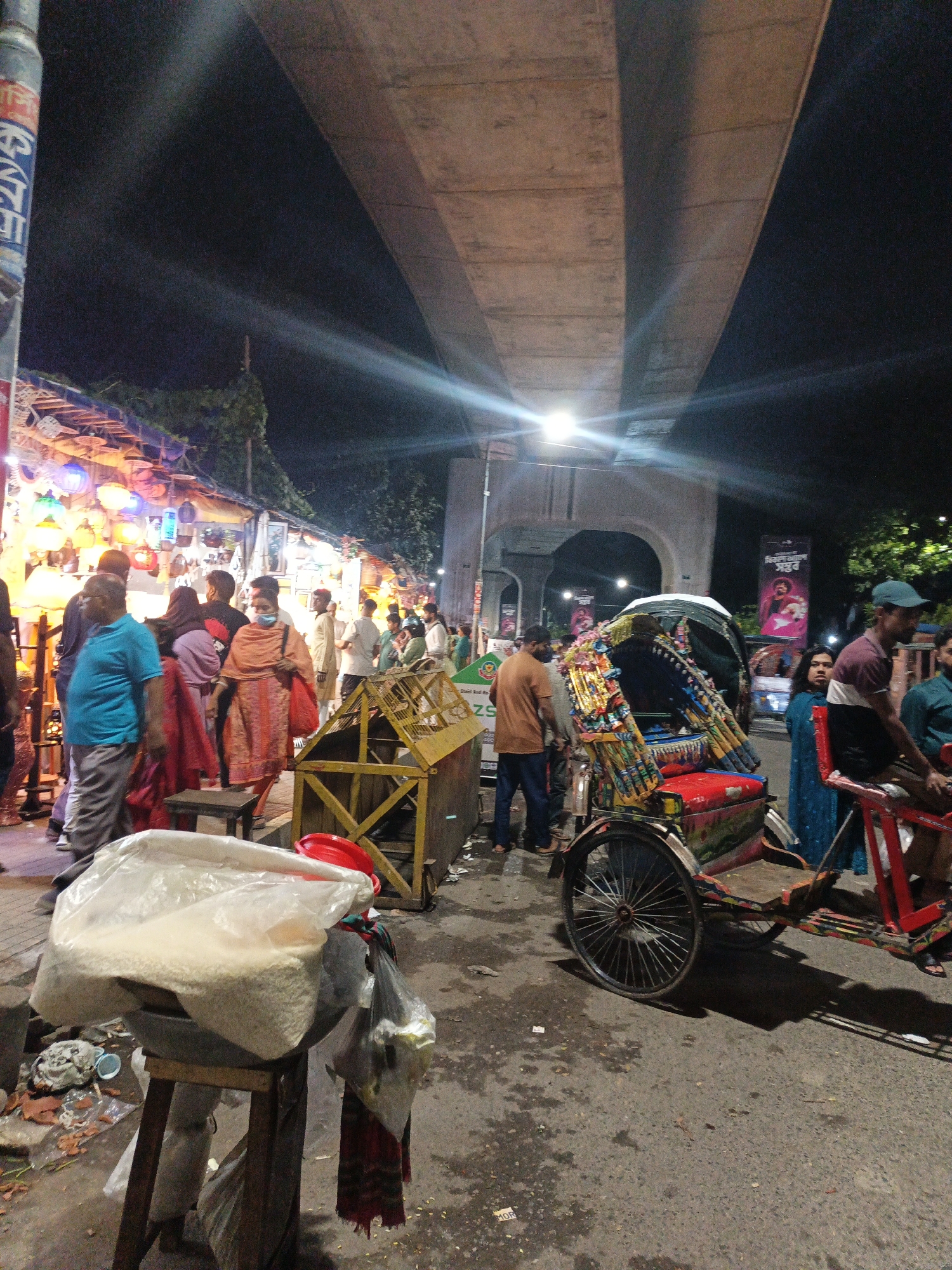 The image depicts a street scene at night in Dhaka, Bangladesh, likely under an overpass or flyover, evidenced by the concrete structure overhead. The street is paved and appears somewhat worn. On the left, a market stall is set up with numerous colorful lanterns hanging, illuminated by artificial light, creating a vibrant display. Several people are browsing or walking past the stall.
In the mid-ground, a decorated cycle rickshaw, a common mode of transport in Dhaka, is prominently featured. It is brightly adorned with painted designs and fabric, with a red seat and a canopy. A few individuals are standing around the rickshaw.
In the foreground, a large plastic bag filled with white granular material, possibly rice or sugar, sits on a wooden stool. Next to it, a black trash bag is hanging. The ground is littered with debris. The overall atmosphere suggests a bustling market or street vendor area at night, with a mix of commercial activity and pedestrian traffic. The lighting is a mix of streetlights, stall lighting, and possibly light from the vehicles.