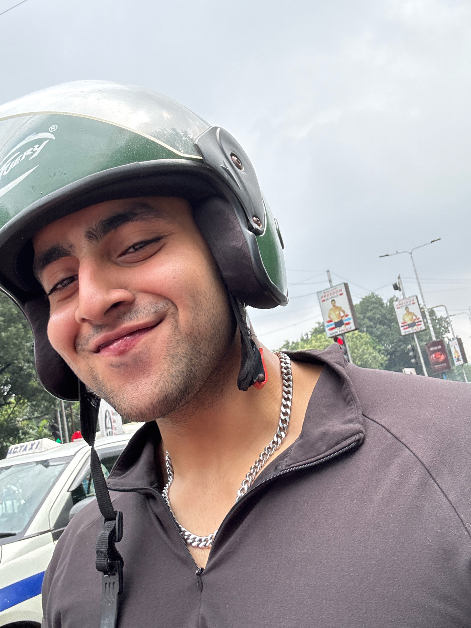 A close-up, selfie-style portrait of a young man wearing a green and silver helmet, a silver cuban link chain, and a dark zip-up jacket. He is smiling widely, with his eyes half-closed. The background suggests an urban environment, with a white taxi visible on the left and billboards on poles in the distance. One of the billboards shows a man in athletic wear with the word "FREEDOM." The sky is overcast. The man is positioned in the foreground, with the rest of the scene blurred, suggesting movement. The overall impression is of a candid shot taken while on the go, likely in Kolkata, India, given the presence of a taxi with "ACTAXI" visible.