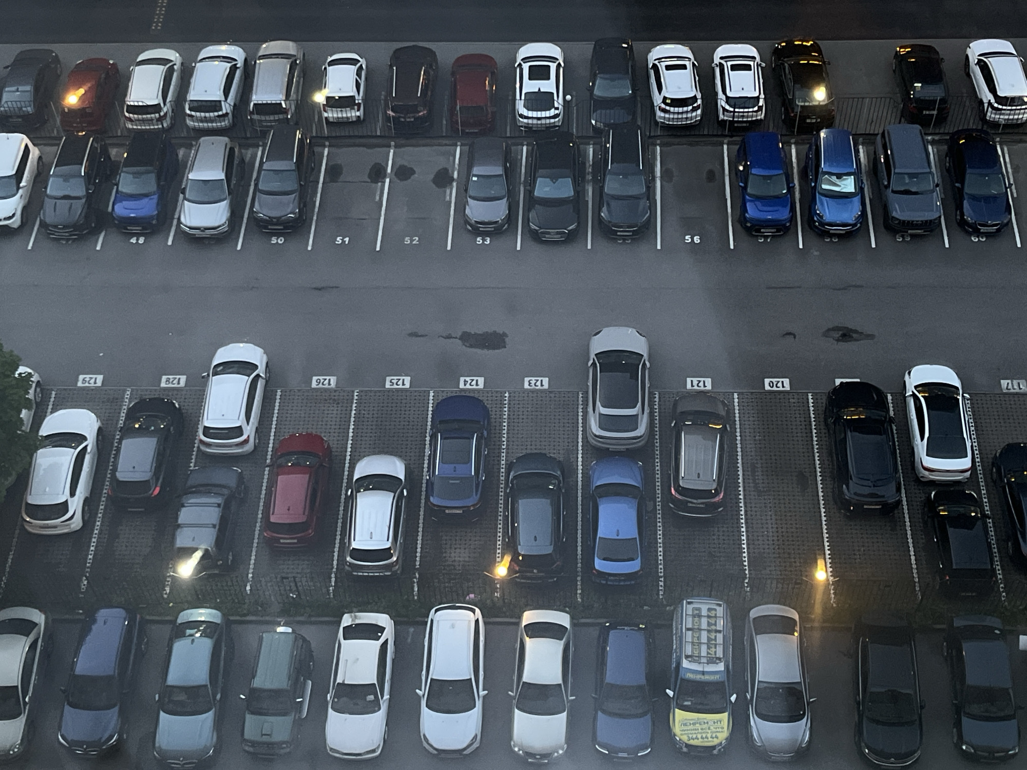This is an aerial, high-angle shot of a parking lot filled with a variety of cars. The parking lot appears to be outdoors, possibly in an urban or suburban area, with a grey, paved surface. The cars are parked in designated spaces marked by white lines, and some spaces have numbers visible, such as 48, 50, 51, 52, 53, 55, 56, 117, 120, 121, 123, 124, 125, 126, 128, and 129.
The cars are of different makes, models, and colors, including silver, black, white, blue, and red. Some cars are parked neatly, while others are slightly askew. There are no people visible in the image. The lighting suggests it could be daytime, but the light is somewhat dim, possibly indicating an overcast day or late afternoon. A few streetlights are visible, some illuminated, suggesting it might be evening or the parking lot is well-lit. One car has its headlights on, casting a bright beam.
There is some visible text on the cars, including what appears to be a yellow service vehicle with text in Cyrillic script, "ЛЕНРЕМ НT", and a phone number "3444444". The overall scene conveys a sense of order and routine, typical of a parking lot. The setting is functional, designed for vehicle storage.