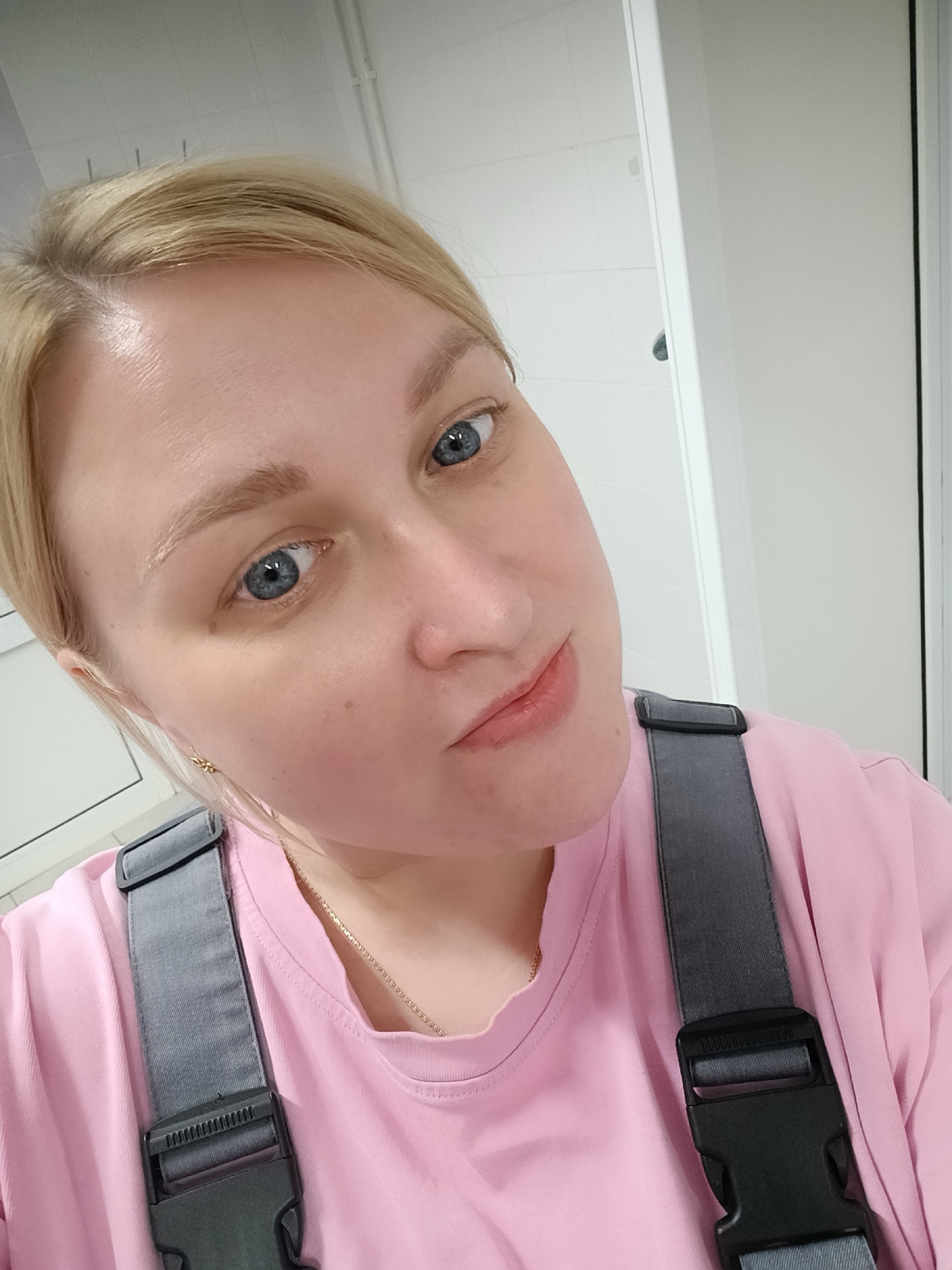 A close-up selfie captures a fair-skinned woman with blonde hair and striking blue eyes, looking directly at the camera with a subtle, pleasant expression. She appears to be in her 20s or 30s.
The woman is wearing a light pink short-sleeved t-shirt, partially covered by grey overalls with visible black plastic buckles and adjustable straps. A thin gold chain necklace is visible around her neck, and a small, intricate gold earring adorns her right earlobe.
The background suggests an indoor, functional setting. Predominantly white walls, possibly tiled, are visible on the left side, while a distinct vertical white doorframe with a dark edge runs along the right side of the frame. Above her head, white ceiling panels and thin, dark linear fixtures (potentially pipes or lighting tracks) are visible. The overall lighting is bright and even, indicative of indoor illumination.
The scene takes place indoors, likely in a commercial, office, or institutional building in Chaikovsky, Russia. No visible text is present.