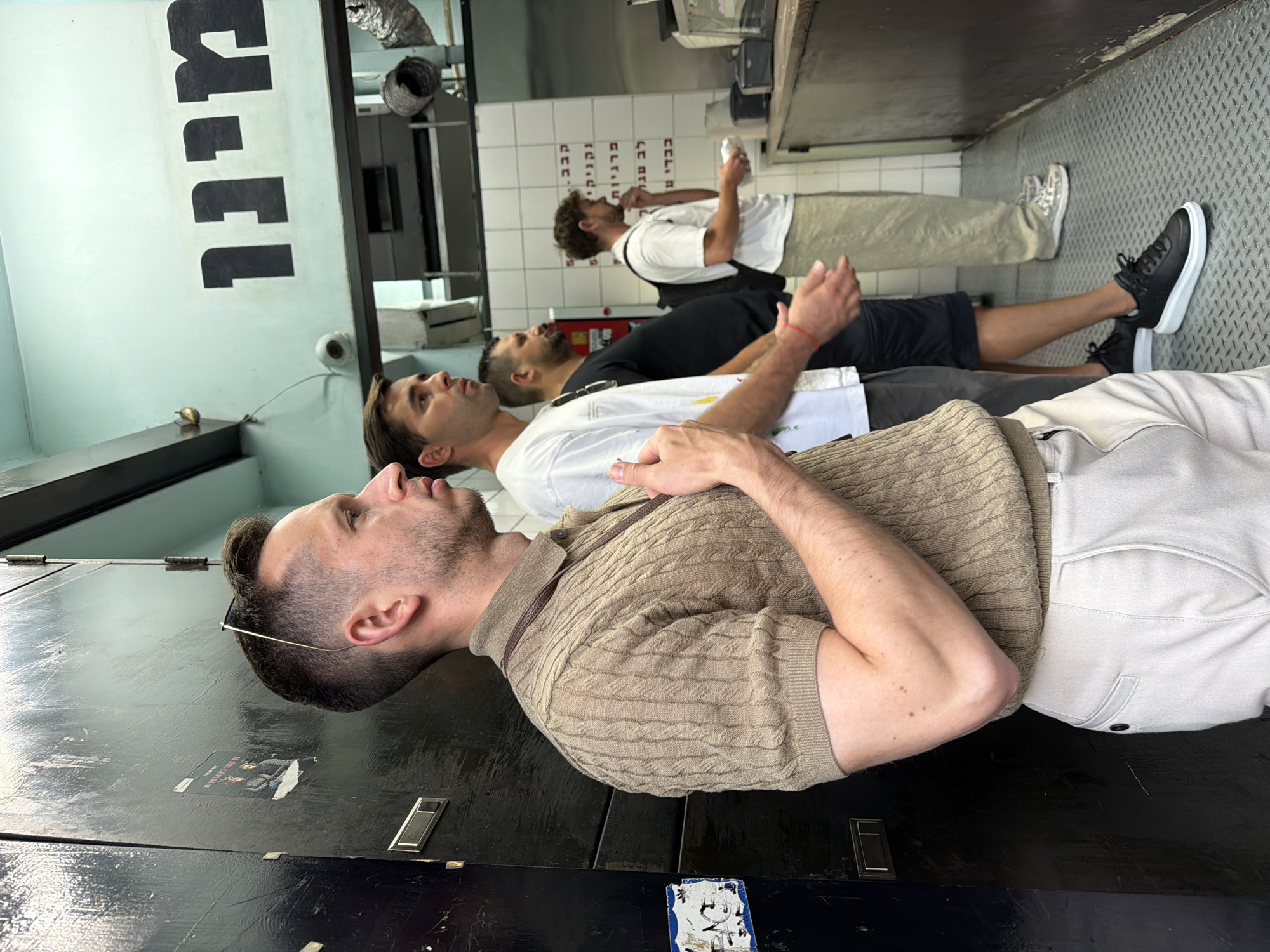 Three men are standing inside a building, likely a shop or restaurant, in Tel-Aviv, Israel. The man in the foreground has a light-brown collared shirt and light-colored pants. Behind him, the second man has a white T-shirt and gray pants. The third man has a black T-shirt and black shorts. All three men are facing the same direction. A fourth man is visible in the background, wearing a white T-shirt and light-colored pants. Hebrew writing is visible on a tiled wall, likely menu items. The floor appears to be made of metal.