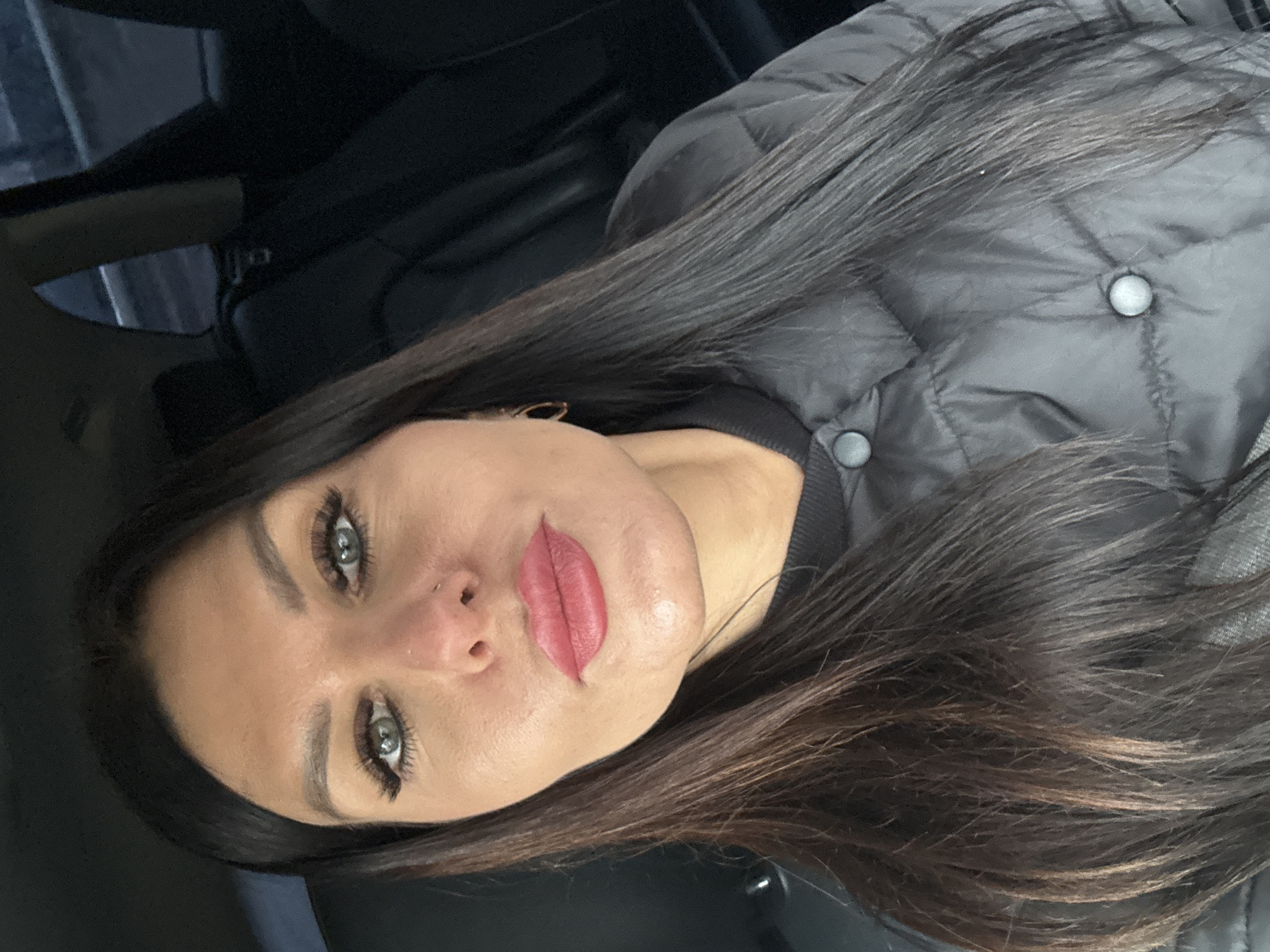 A woman with long, dark hair and light eyes is pictured from a close-up, slightly angled perspective. She is wearing a quilted jacket. The background appears dark, suggesting an interior setting, possibly a car. There is no verifiable information to suggest a location in the city of Chaikovsky, Russia.
