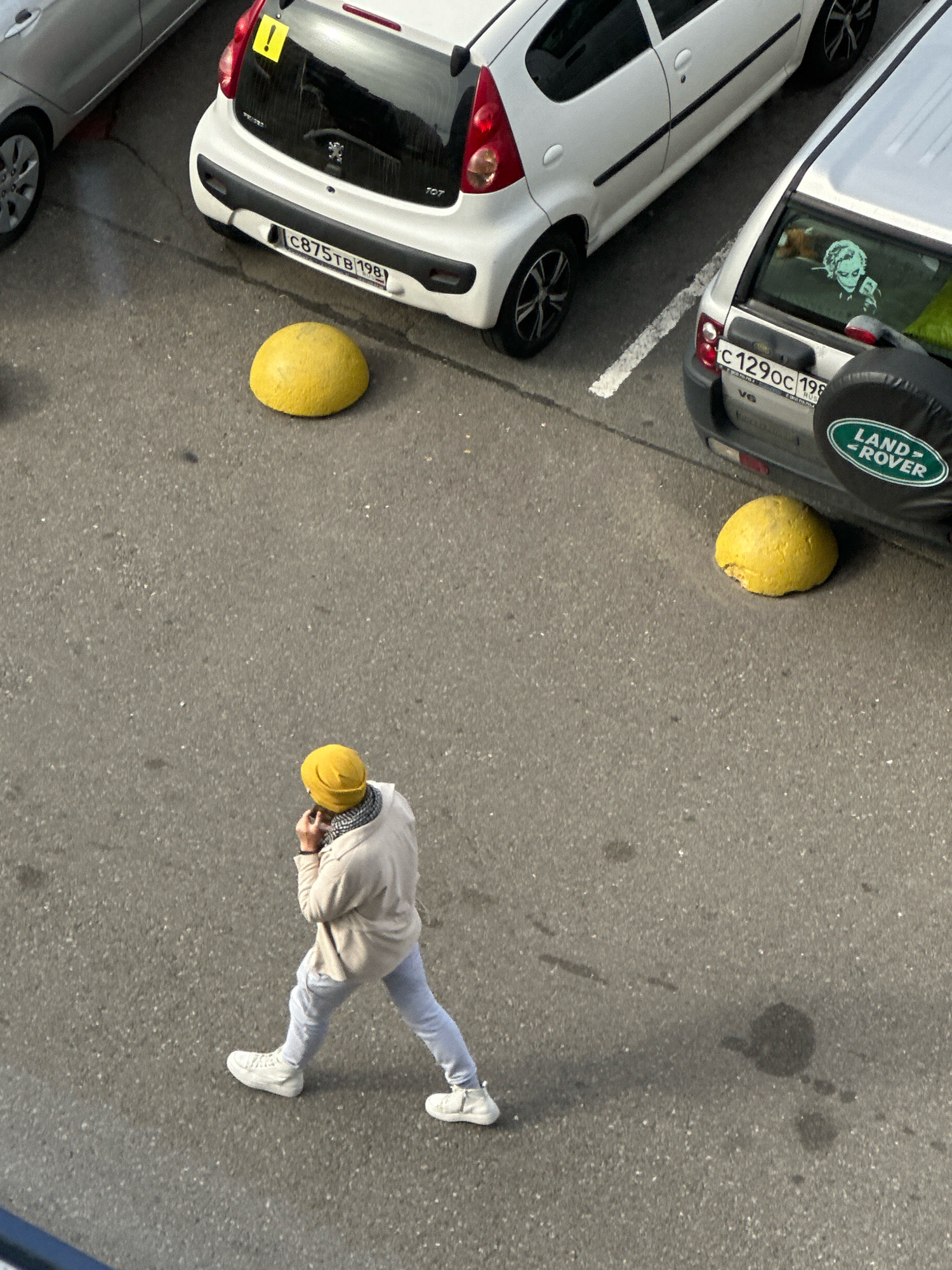 An overhead, full-frame shot captures a street scene in Saint Petersburg, Russia. A person, viewed from above, walks across a gray asphalt surface. They wear a yellow beanie, a light-colored hooded jacket, light gray pants, and white sneakers. The person is holding a phone to their ear. In the background, two cars are parked: a white hatchback and a silver SUV. The hatchback has a yellow exclamation mark sticker on its rear window and a license plate that reads "C875TB 198". The SUV has a spare tire cover with a "LAND ROVER" logo and a license plate that reads "C129OC 198". Two yellow, dome-shaped concrete bollards are present in the scene, one in front of each car, designed to prevent vehicles from parking. The asphalt surface shows some dark oil stains and is speckled with small white particles. The lighting suggests it is daytime, with shadows cast on the ground.