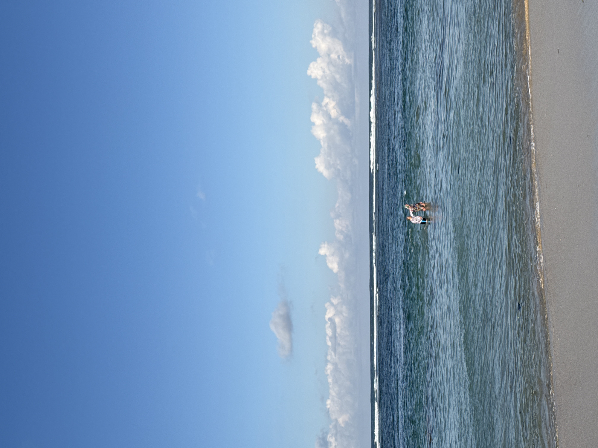 This image presents a serene beach scene, captured from an elevated perspective looking down upon the ocean and shore. The upper half of the frame is dominated by a clear, bright blue sky dotted with scattered white cumulus clouds, some clustered near the horizon line and others appearing isolated.
The ocean occupies the central part of the image, displaying clear water with a gradient of blue and greenish hues. Gentle waves are visible, especially closer to the shore, where white foam patterns trace the waterline.
To the right, the light beige sand of the beach meets the ocean. The texture of the sand, wet near the water and drier further up, is discernible.
Two individuals are visible in the shallow ocean water towards the bottom center. One person, wearing a dark patterned swimsuit, is wading or partially floating, seemingly holding a small, light-colored object, possibly a boogie board or float. Another person is partially obscured beside them, also in the water. Their presence suggests leisurely activity, enjoying the ocean.
The scene is brightly illuminated, indicating a clear, sunny day, likely mid-day. The weather appears perfect for beach activities. The tranquil atmosphere and natural elements are consistent with a tropical coastal setting like Kuta Selatan, Indonesia. No visible text is present in the media file.