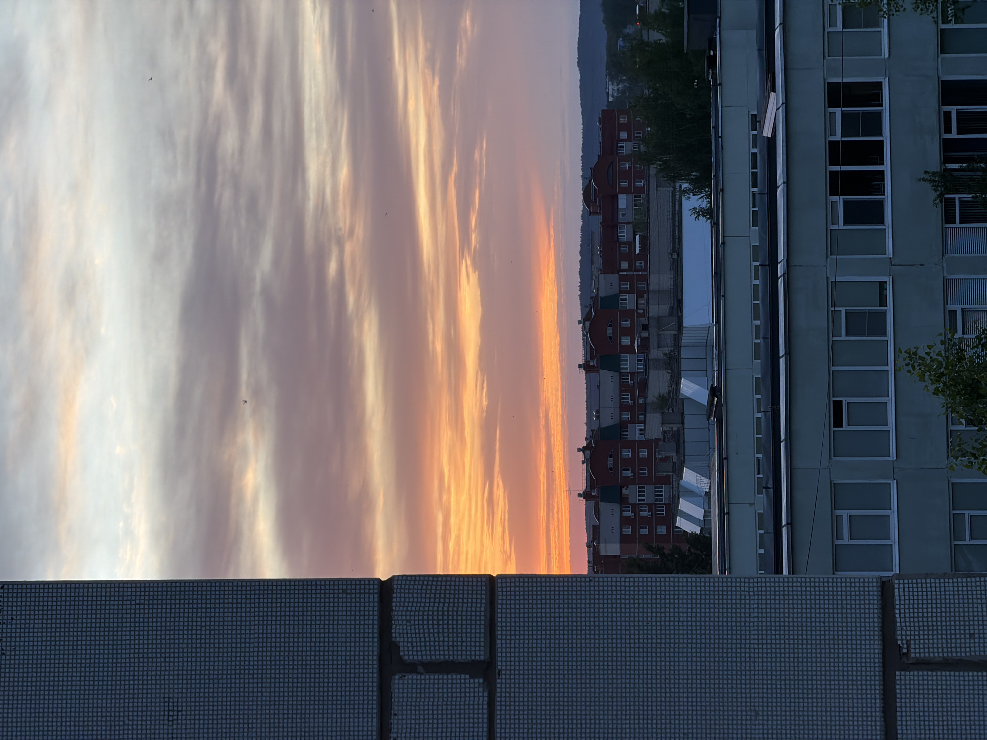 The image shows a sunset view over a city landscape, likely in Chaikovsky, Russia. In the foreground, there is a low wall covered in blue-gray square tiles. The cityscape includes red and gray apartment buildings in the mid-ground, and a light blue-gray building with multiple windows of varying sizes on the right side. The sky is filled with dramatic clouds reflecting orange and yellow hues from the sunset. The horizon is visible behind the buildings, with a hint of trees or a hill in the distance.