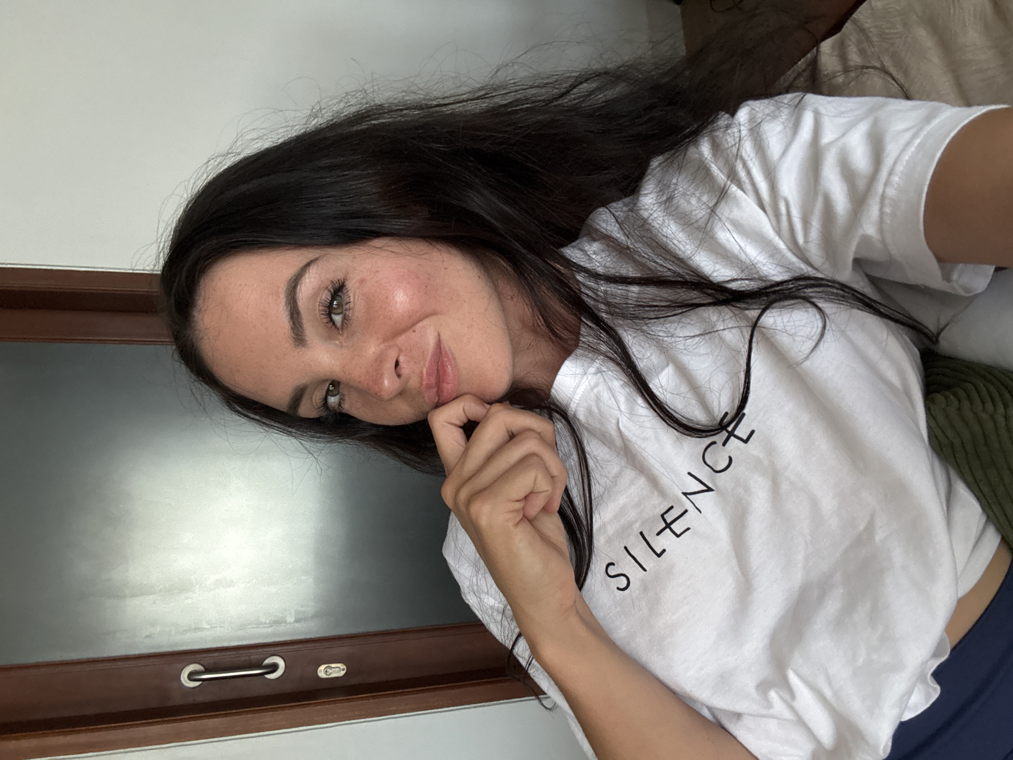 A young woman with long, dark hair and fair skin is captured in a close-up, self-taken photograph. She is wearing a white t-shirt with the word "SILENCE" printed in black, block letters across the chest. Her right hand is raised, with her fingers gently touching her chin, and she is looking towards the camera with a slight smile. She has green eyes and freckles across her nose and cheeks. The background appears to be an indoor setting, possibly a bedroom, with a wooden doorframe and a grey, textured surface visible to the left, and a plain white wall to the right. A hint of blue fabric, likely part of her lower garment, is visible at the bottom of the frame. The lighting suggests it is daytime, possibly near a window. The image does not contain any specific landmarks or visual cues that would definitively place it within North Kuta, Indonesia.