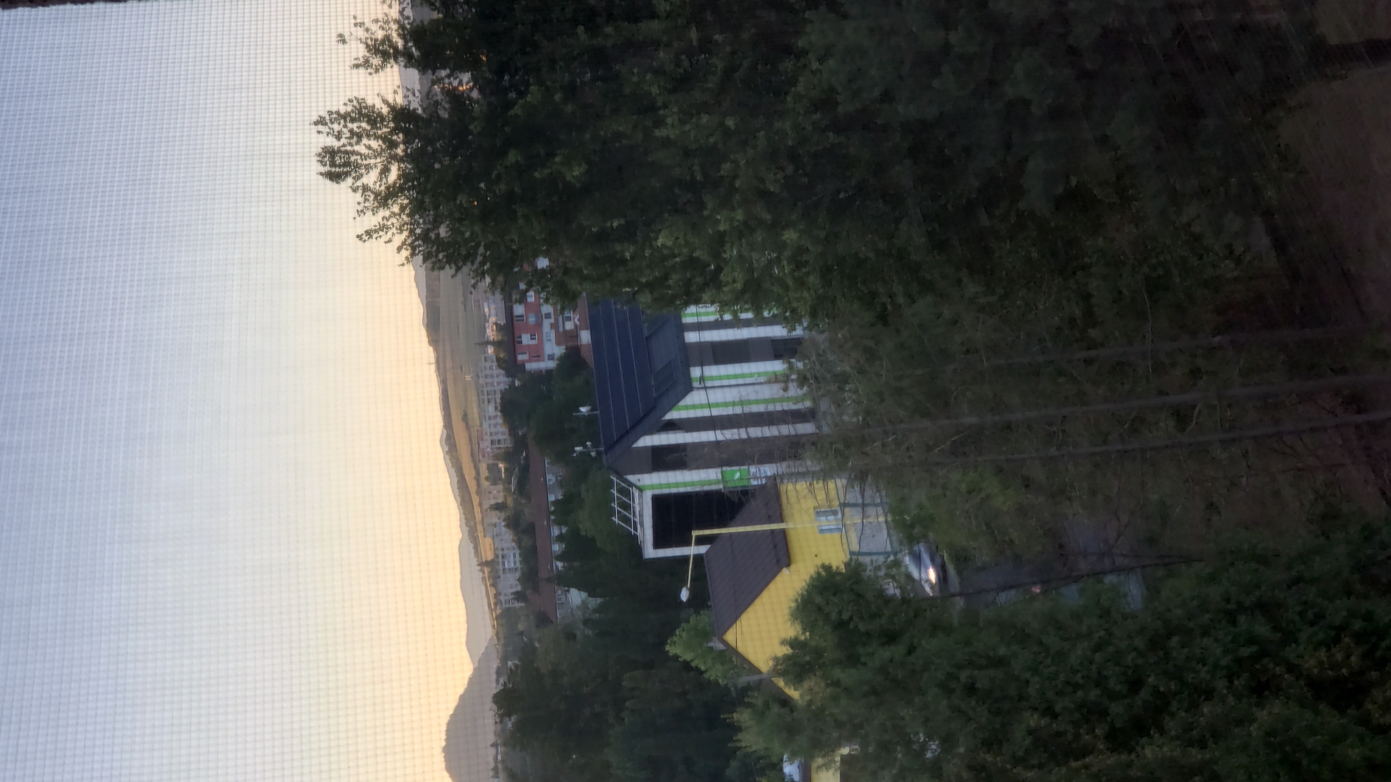The image shows a view of a residential area in Prešov, Slovakia, with a street and buildings visible. The foreground is dominated by trees, some of which are quite dense. A yellow house with a dark roof is prominent on the left side of the image, with a modern building featuring a dark roof with solar panels and vertical white and green stripes situated next to it. A streetlamp is positioned between these two structures. Further in the background, there are more buildings and residential blocks, suggesting a neighborhood. The landscape beyond the immediate buildings includes hills and mountains under a hazy sky, with the soft light of either sunrise or sunset casting a warm glow on the distant horizon. A silver car is partially visible on a road in the middle ground, indicating a functioning urban environment. The overall scene is captured through a mesh screen, which creates a grid pattern across the entire image, slightly obscuring the details.