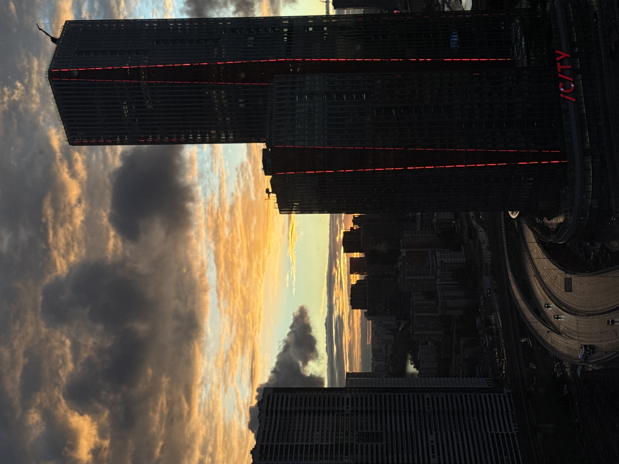 The image captures a cityscape at sunset or sunrise in Khimki, Russia. Prominently featured is a modern, dark-colored skyscraper with horizontal red illuminated stripes and the letters "iCITY" displayed vertically on one side. Below the skyscraper, there's a view of other buildings, city infrastructure, and a curved highway with moving vehicles. The sky is filled with dramatic clouds colored with hues of orange, yellow, and gray due to the setting sun or rising sun, creating a striking contrast between the natural and urban elements of the scene.