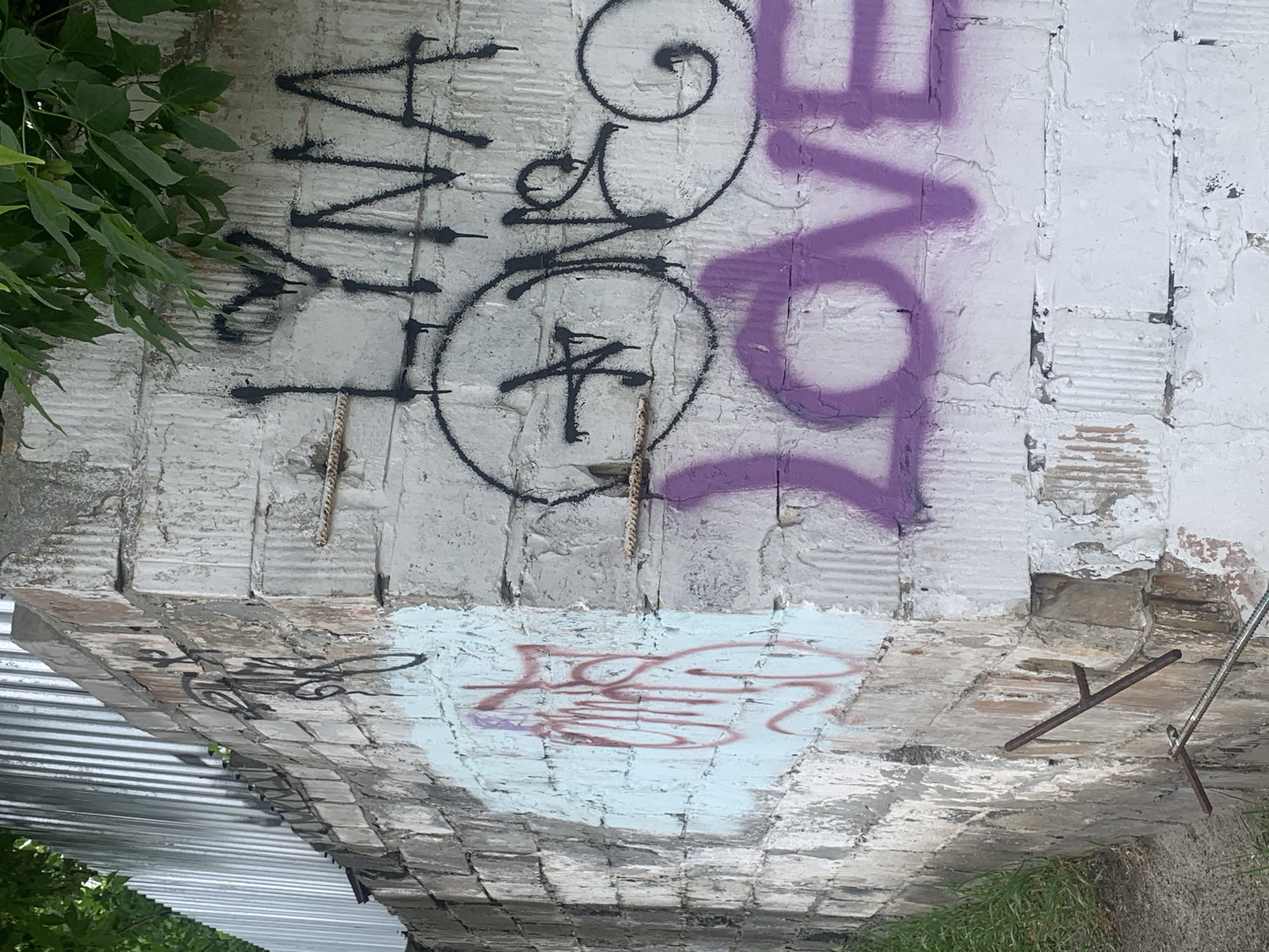 The image shows a concrete and brick structure in Kyiv, Ukraine, adorned with graffiti. A partial view of a corrugated metal roof and greenery are visible. The structure has been painted white and is covered in various graffiti designs, including the word "love" in purple paint, and other black painted symbols and writing. The structure shows signs of decay with exposed brick and rebar.