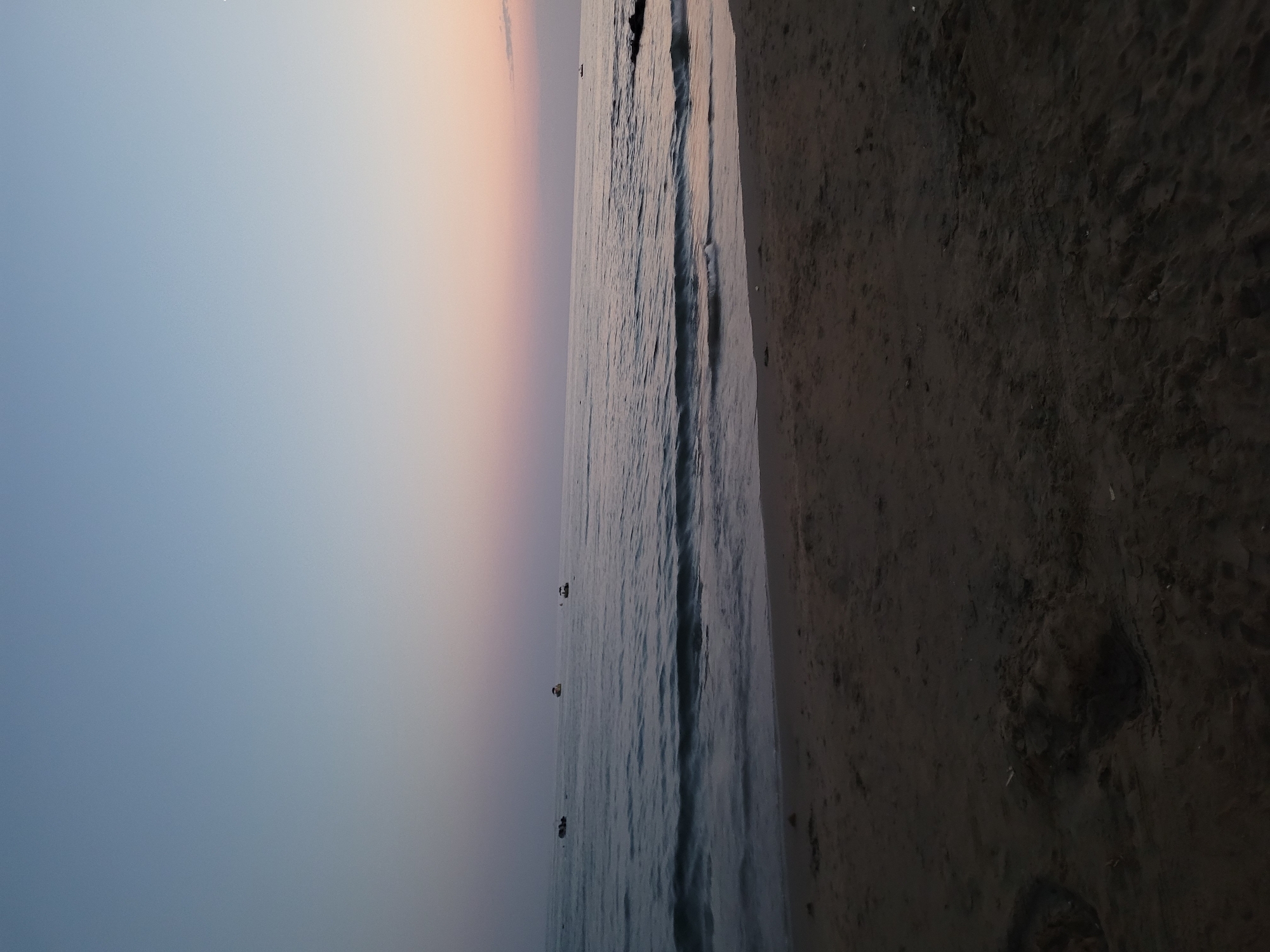 The image depicts a tranquil beach scene at dusk in Manavgat, Turkey. The sky transitions from a pale blue at the top to soft hues of pink and peach near the horizon, indicating the end of daylight. The calm sea reflects the sky's colors, with gentle waves lapping at the sandy shore. In the distance, three small boats are visible on the water. The foreground is dominated by the dark sand of the beach, which appears smooth and undisturbed. The overall atmosphere is peaceful and serene, characteristic of a sunset by the sea.