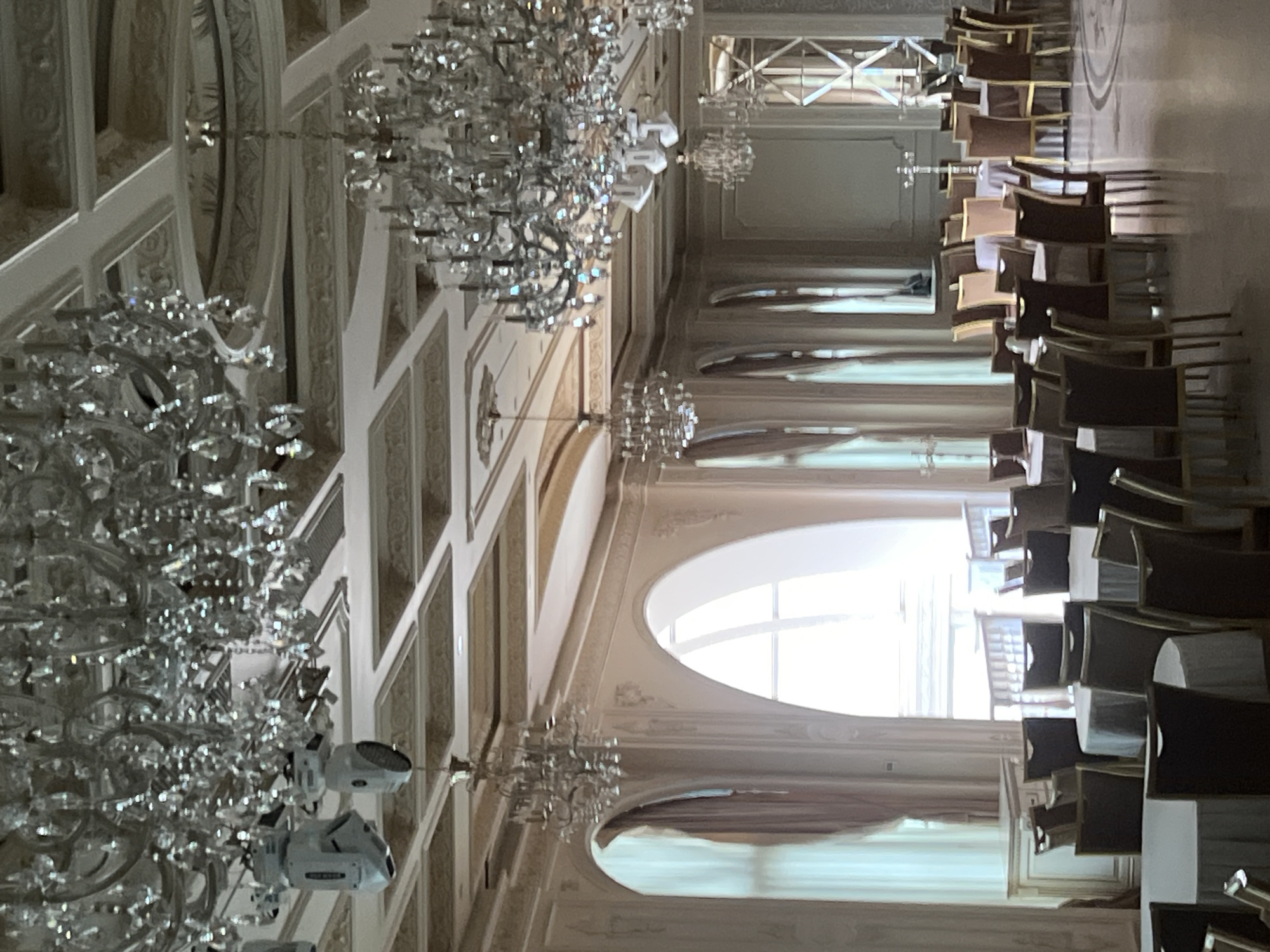 The image displays a grand and elegantly decorated hall, likely a ballroom or event space, set up for a formal gathering. The perspective is from an elevated position, looking across the length of the room.
The most prominent features are the numerous elaborate crystal chandeliers that hang from a very high ceiling. The ceiling and upper walls are adorned with intricate white and cream-colored decorative moldings, panels, and archways, contributing to a sense of opulence.
The hall is arranged with many rows of chairs, suggesting it is prepared for an event such as a conference, banquet, or ceremony. The chairs have dark upholstery and light-colored or gold frames, neatly aligned in multiple columns that recede into the distance. No people are visible in the room, indicating it is either before or after an event.
One side of the room features large arched windows that allow abundant natural light to flood the space, suggesting it is daytime. Light-colored curtains or drapes are visible near these windows. The floor appears to be a light-toned surface.
Interspersed among the traditional crystal chandeliers are several modern stage lighting fixtures, one of which has the partial text "SHOW" visible on its casing, implying contemporary event production capabilities within the classic setting. The overall impression is one of grandeur, sophistication, and readiness for a large-scale event in an upscale venue in Yerevan, Armenia.