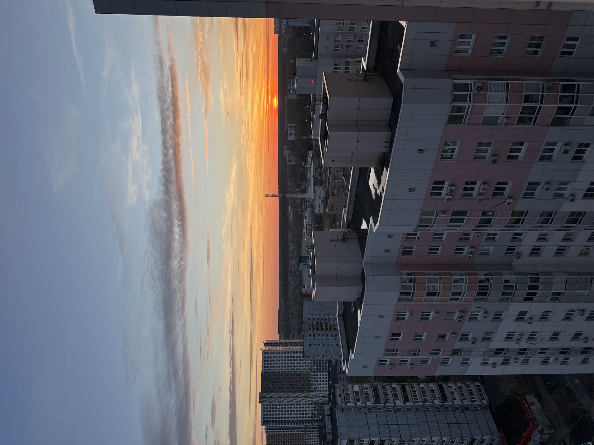 The image depicts a sunset scene over a densely populated urban area. The sky is filled with clouds, exhibiting hues of orange, yellow, and gray. The sun is visible on the horizon, partially obscured by buildings and trees. The cityscape is composed of numerous high-rise buildings. The buildings are primarily gray, with some having pink and white accents. The scene is in Moscow, Russia.