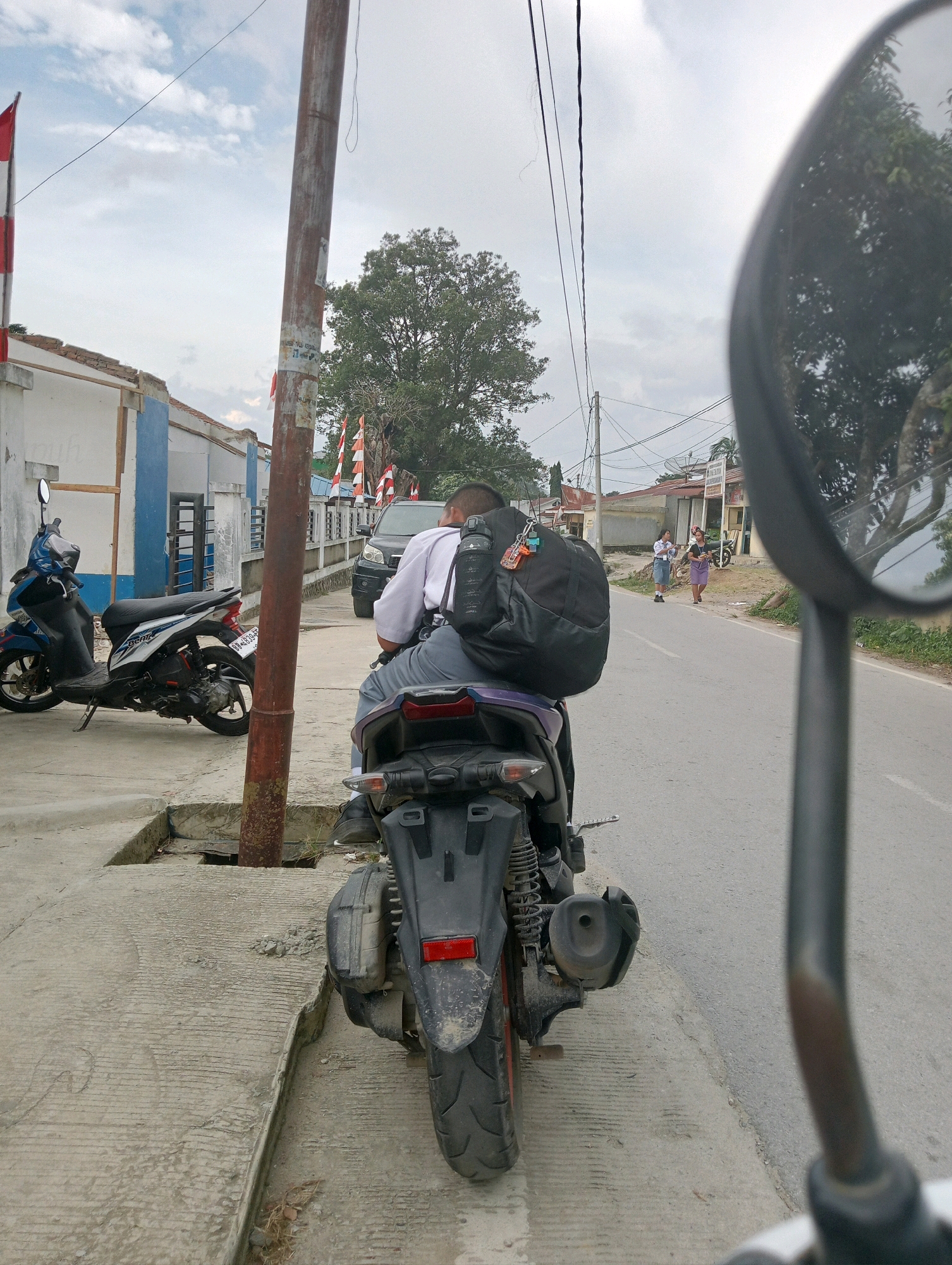 The image captures a street scene in Hinalangbagasan, Indonesia. In the foreground, a person is seated on a black and purple scooter, facing away from the camera. This individual is wearing a white collared shirt and grey pants, typical school attire, and a large black backpack is strapped to their back. The scooter is parked on a concrete sidewalk next to a street. To the left of the scooter, another blue and black scooter is parked.
Further down the street, a black SUV is visible. On the right side of the street, two young people, likely students in uniform, are walking. The environment includes a utility pole with wires running across the scene, trees, and buildings in the background. The sky is overcast with clouds. The reflection of trees is visible in the side mirror of a vehicle in the foreground, suggesting the photo was taken from another vehicle.