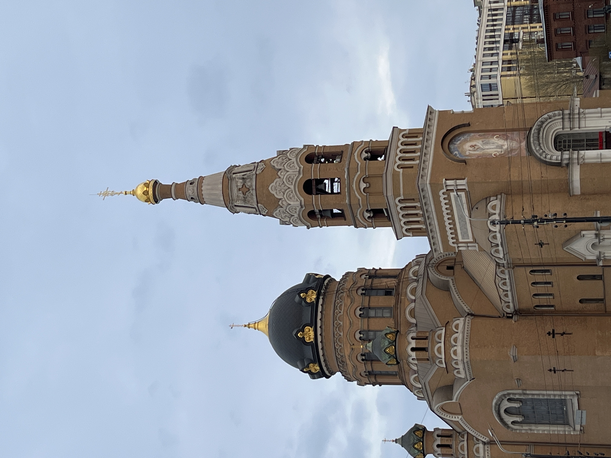 The image shows a tall, ornately designed church in an urban environment. The church is primarily brown with white and gold accents. It has multiple towers, including a prominent one with a pointed spire. Some towers have rounded domes. The facade features arched windows and decorative detailing. In the background, modern buildings are visible, indicating a city location. Electrical wires are strung across the front of the church, further suggesting an urban setting. The sky is overcast with a gray-blue tone. The location is Unknown, Unknown.