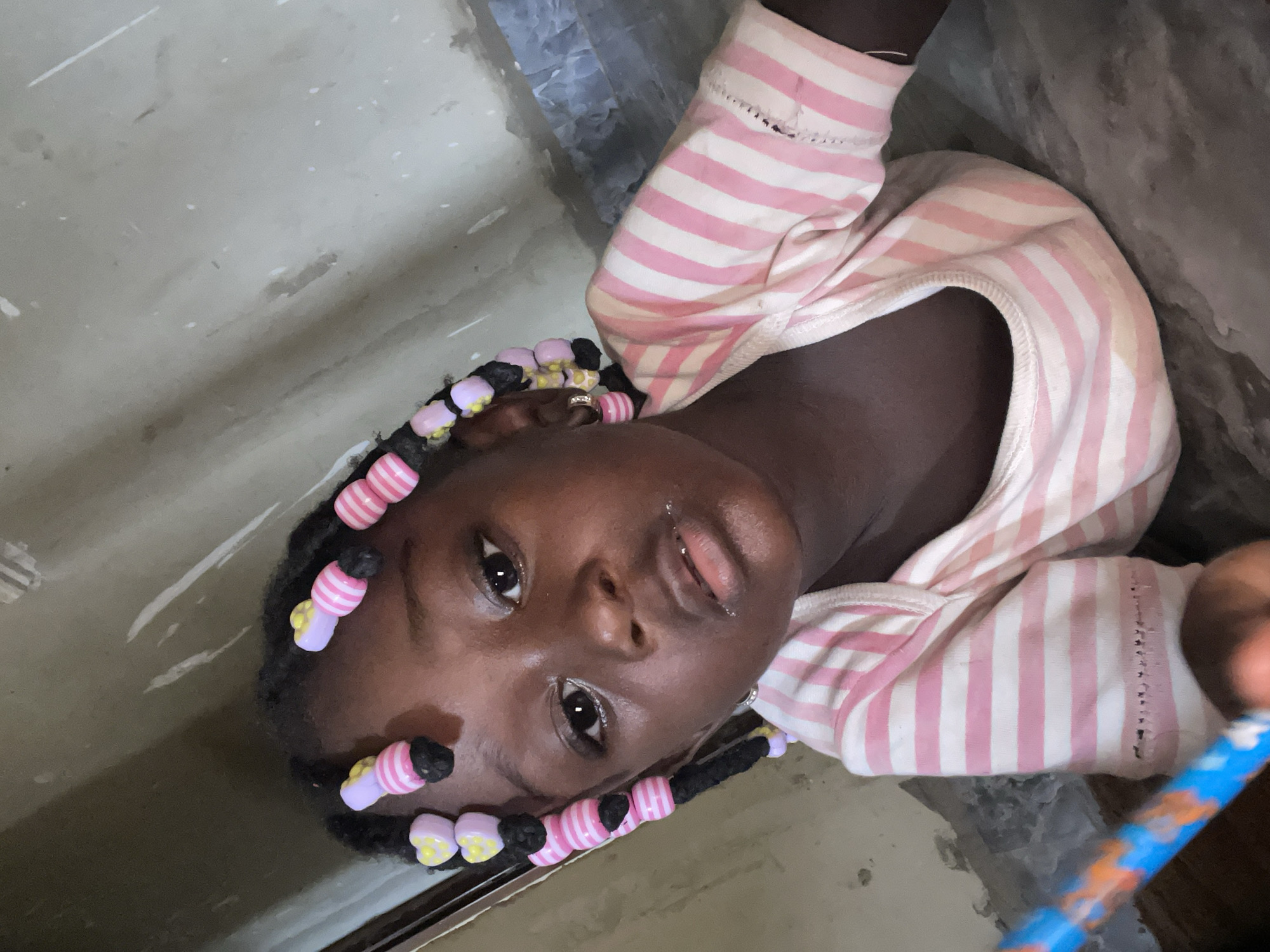 A young Black girl is prominently featured, looking directly at the camera. She appears to be lying down or leaning back, with the image captured at a tilted angle. She wears a long-sleeved top with alternating pink and white horizontal stripes. Her hair is styled in braids, adorned with numerous colorful beads in shades of pink, white, yellow, and light purple, some shaped like hearts or flowers. Her expression is neutral and calm, with a slight glisten on her lips.
The background is a plain, light gray or beige surface, possibly a wall or floor, showing some minor scuff marks or texture. A blue object with orange patterns, resembling a pencil or stick, is partially visible in the bottom right corner of the frame. One of the cropped images shows a small, possibly metallic ring on one of her fingers. The setting appears to be indoors in Akwanga, Nigeria, as per the provided context. There is no discernible text visible in the image.