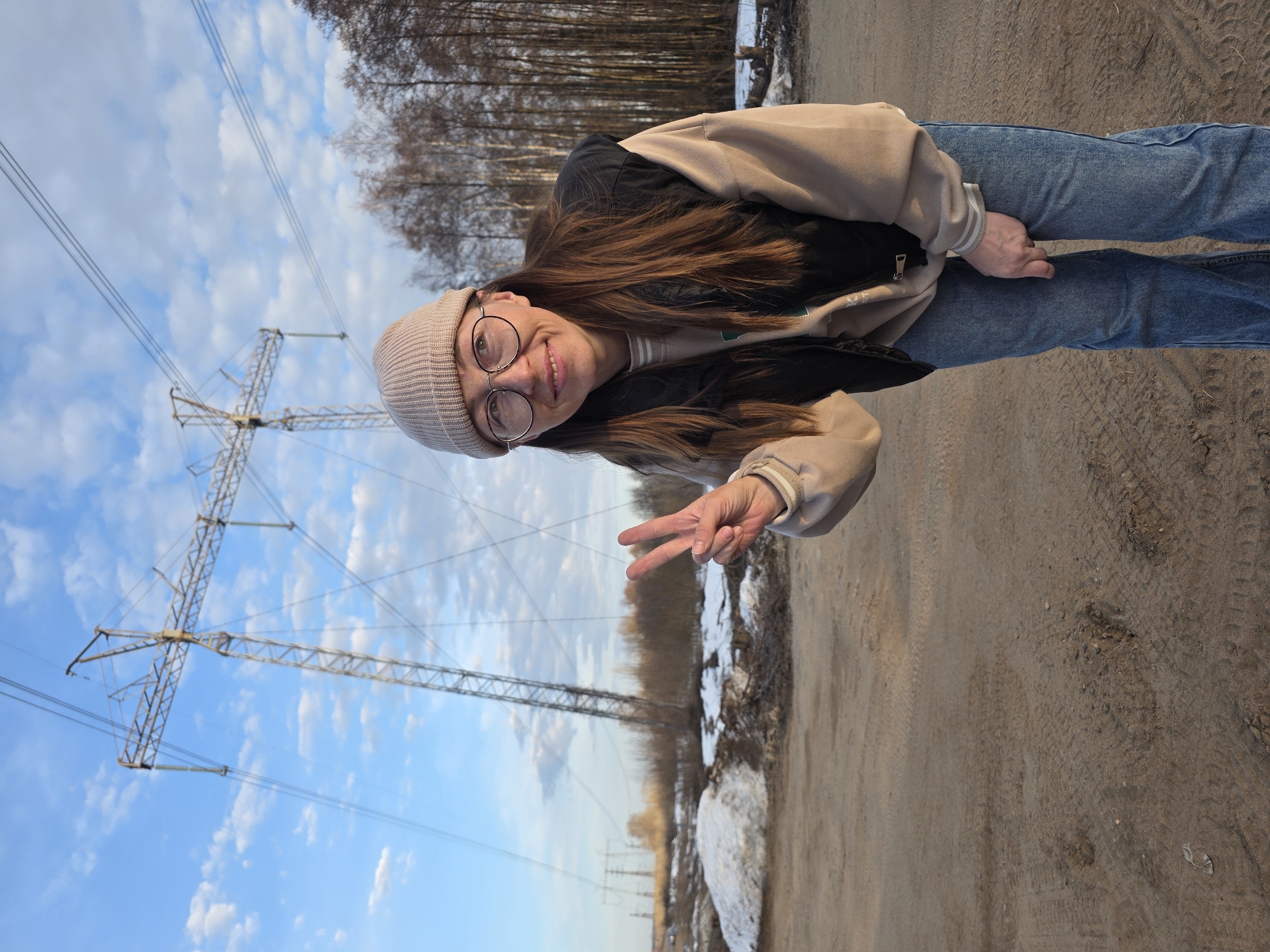 A young woman, wearing a light tan beanie, round glasses, a beige and black jacket, and blue jeans, is standing on a dirt path in Noviy, Russia. She is giving a peace sign with her right hand. Behind her, there's a line of leafless trees and scattered snow. A power line tower is visible to her left against a blue sky with scattered clouds.