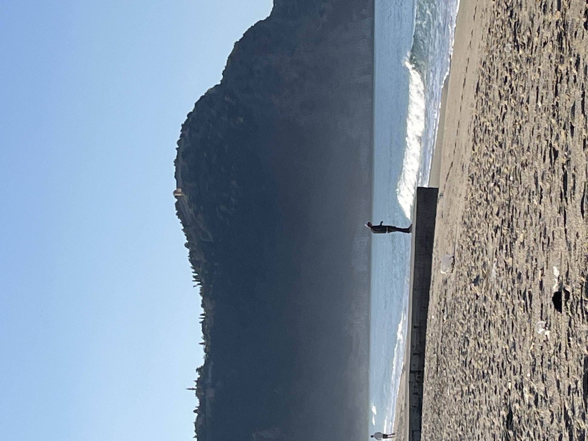 The image depicts a coastal scene in Alanya, Turkey. The left side of the frame features a large, dark, tree-covered mountain. To the right of the mountain is a body of water, likely the Mediterranean Sea, with a visible wave. The shoreline consists of a sandy beach with a dark color. A concrete structure runs parallel to the shoreline, separating the beach from the water.
Two people are visible in the scene. One person stands on the concrete structure, facing the sea. Another person is located on the beach near the water's edge. The sky is clear and blue, suggesting a sunny day.