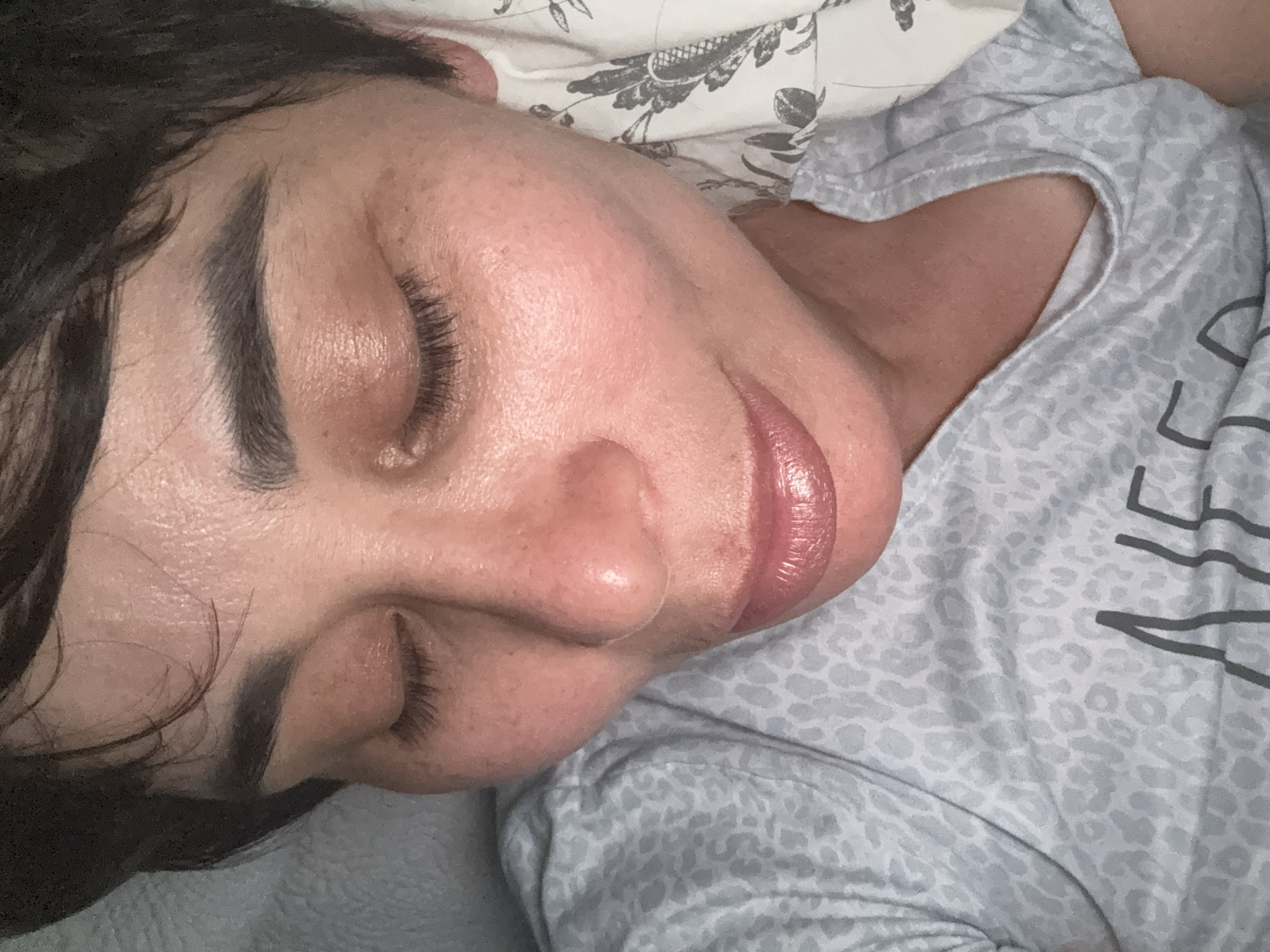 A close-up, overhead shot shows a woman with her eyes closed, lying on her side on a bed with a patterned pillow. She has short, dark hair, dark eyebrows, and appears to be wearing light-colored makeup, including lipstick. Her skin has a slightly flushed appearance and some texture. She is wearing a light gray t-shirt with a subtle animal print and appears to be indoors. The background consists of a textured gray surface and the floral patterned pillow. There is no indication of the city of Bellmore, United States of America.
