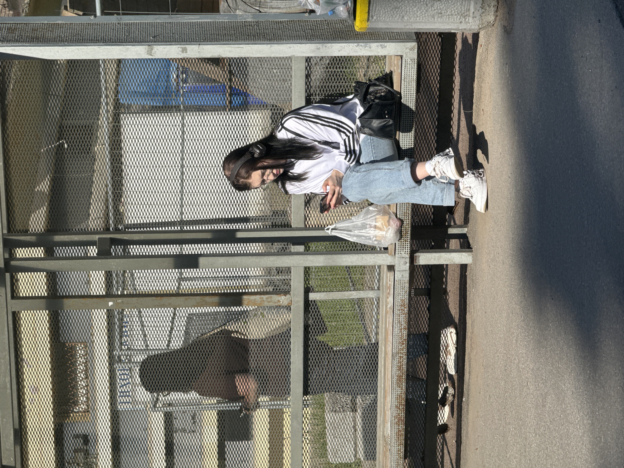 A young woman is depicted sitting on a metal bench or ledge outdoors, seemingly resting or waiting. She is looking downwards, holding a smartphone in her hands. Her long, dark hair falls over her shoulders, and she wears a white long-sleeved top with three black stripes on each sleeve, light blue jeans, and white sneakers with white socks. Black over-ear headphones are visible around her neck.
Beside her on the metal surface is a black bag, possibly a handbag or backpack. A clear plastic bag containing what appears to be food items rests on the ground near her feet. The immediate surroundings are dominated by a sturdy metal structure, possibly a fence, barrier, or part of a public shelter, made of dark grey frames and a diamond-patterned mesh.
The scene is brightly lit by natural sunlight, casting strong, distinct shadows on the asphalt or paved surface to the right of the woman and the metal structure, suggesting it is daytime, possibly late morning or afternoon. Through the mesh of the structure, glimpses of a blurred background can be seen, including hints of blue and green. A faint, blurred reflection of a person, likely the photographer, is visible within the mesh panels in the lower left portion of the image. No visible text can be clearly discerned in the media.