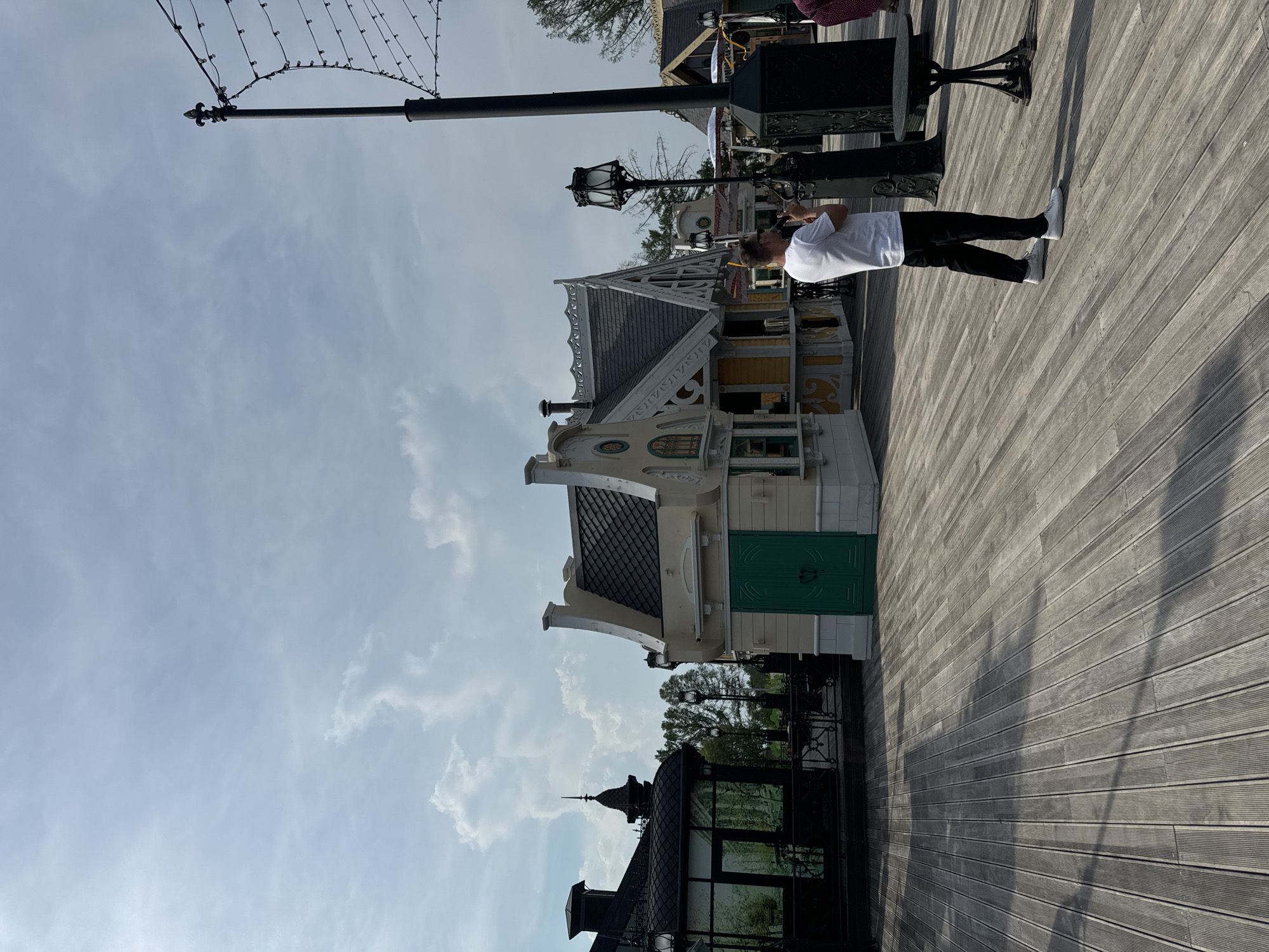 The image depicts a person standing on a wooden boardwalk, holding a phone and seemingly taking a picture. Behind the person are several small, whimsical buildings with decorative gables, resembling stalls or small shops. One building features a bright green door. To the left, a dark-colored building with glass panels and an ornate spire is visible. A tall lamppost with string lights is present on the right side of the boardwalk, along with a small table and chairs. The sky above is partly cloudy, with a bright sun casting shadows on the boardwalk. The overall scene suggests an outdoor leisure or entertainment area, possibly a park or amusement area. There is no indication of the specific location within Moscow, Russia.