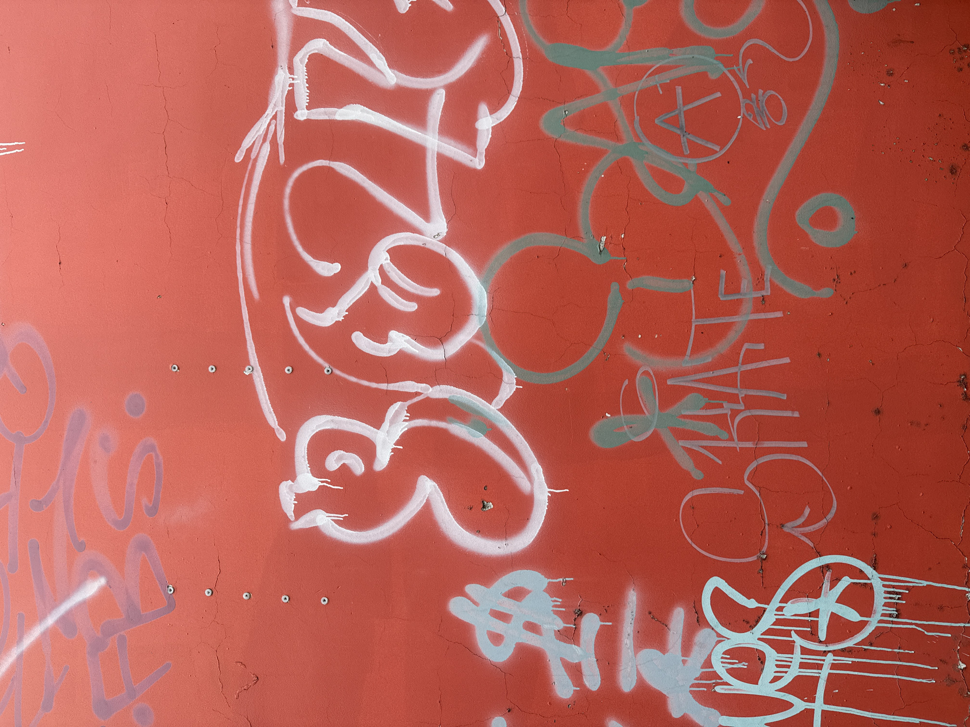 The image displays a section of a red, textured wall covered in graffiti. The graffiti is rendered in various colors, including white, light blue, and a greenish-gray. There are prominent spray-painted lettering and abstract shapes. Some of the lettering appears to be stylized words or tags. The wall itself shows signs of age and weathering, with visible cracks and scattered debris. There are no people or animals present in the image. The context of Da Lat, Vietnam, is not evident from the visual information provided in the image.