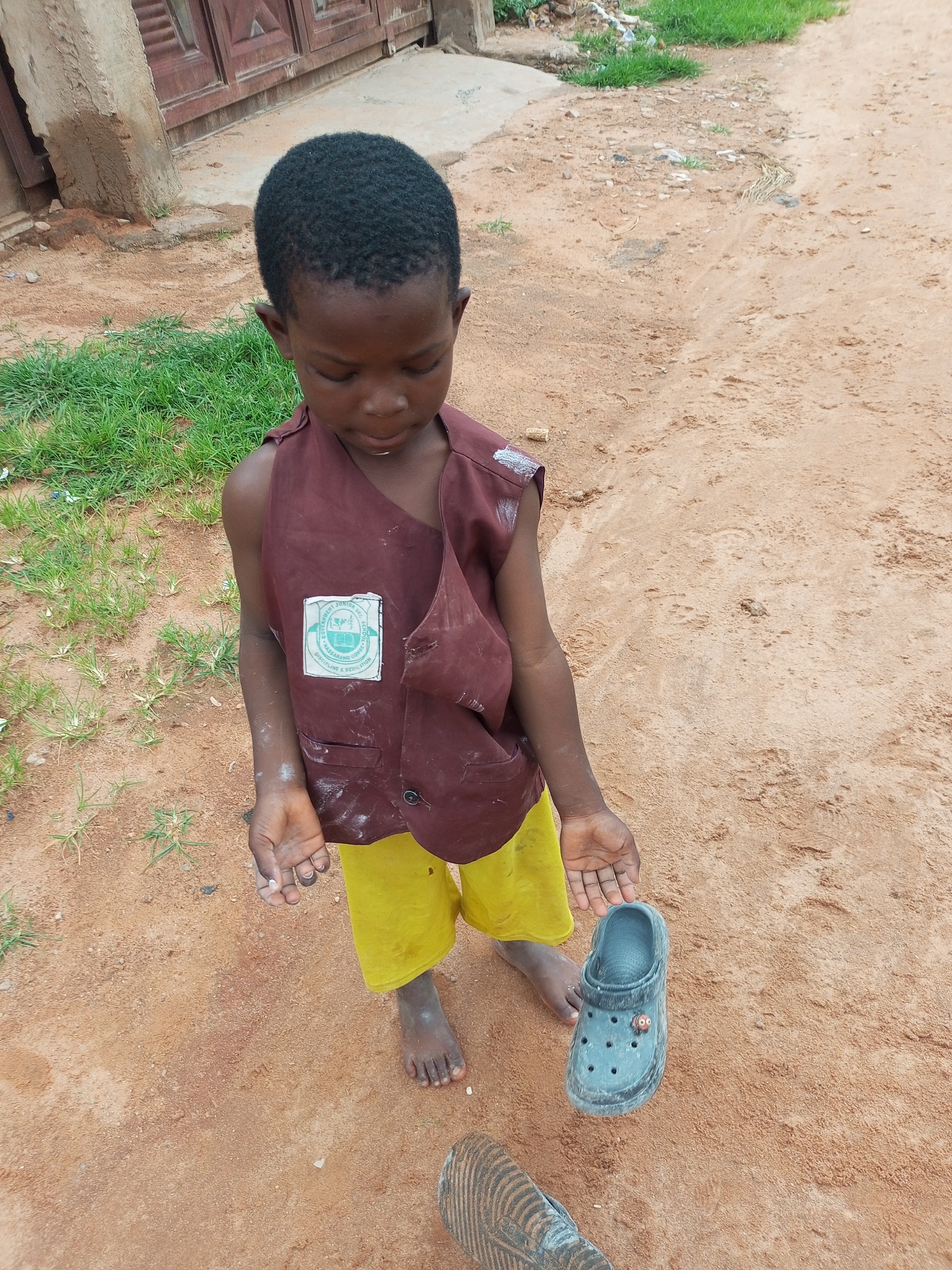 A young boy stands on a dirt path in a village setting, possibly in Tempure, Nigeria. He wears a maroon vest over yellow shorts and is barefoot. The vest has a circular emblem with text and imagery, suggesting a school uniform. The boy is holding a single blue Croc shoe in his right hand and looking down at it. His left hand is open and held slightly away from his body. The ground is sandy and dry, with patches of green grass and some debris visible in the background. A building with wooden doors is present behind him. The overall lighting suggests an outdoor, daytime scene.