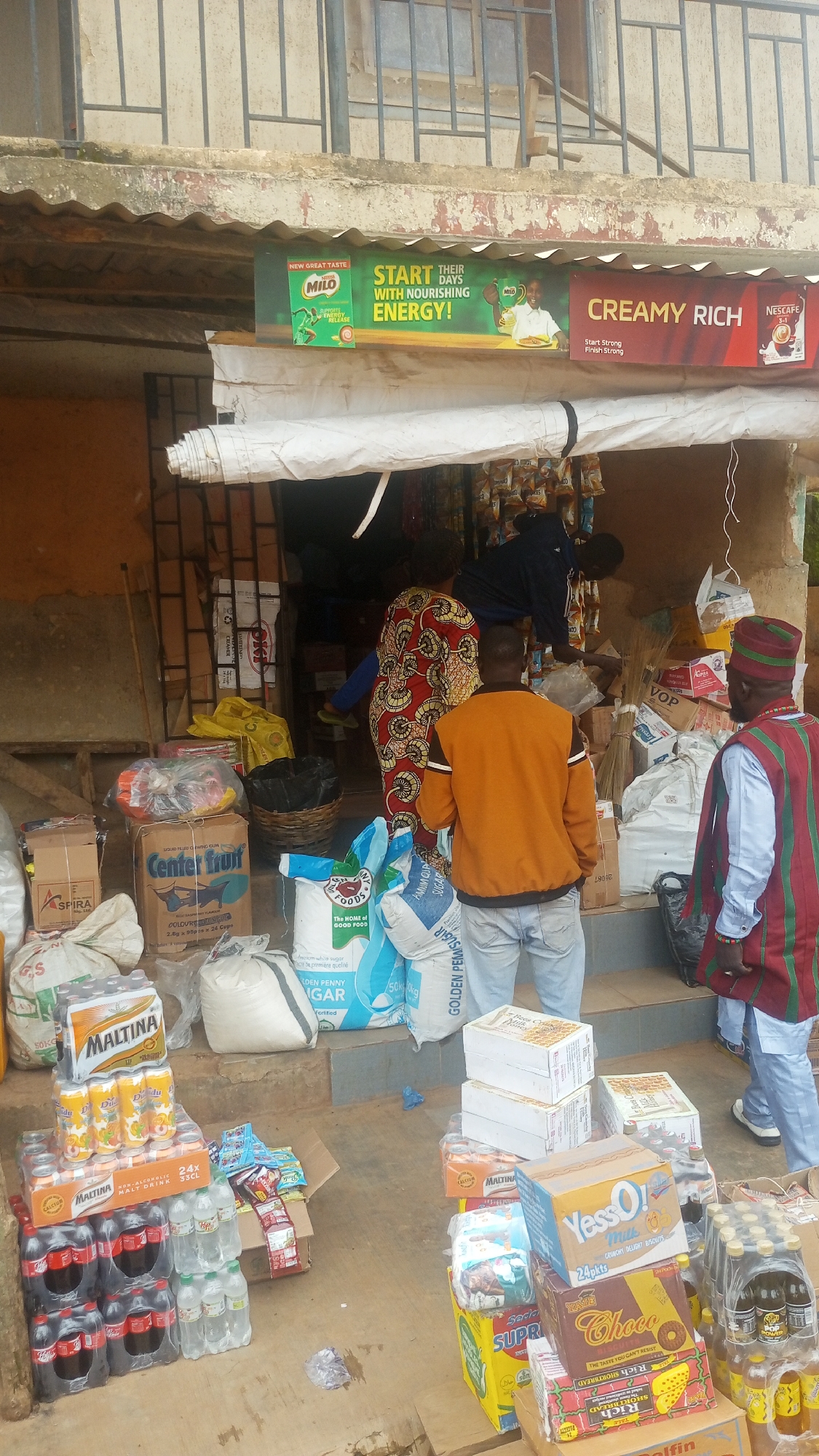 A street-level view captures a small shopfront in Bukuru, Nigeria, with a modest display of goods for sale. The shop is open to the street, with products stacked both inside and on the ground in front of it. A corrugated metal awning extends over the entrance, where several advertisements are displayed. One prominent ad promotes Milo, encouraging consumers to "START THEIR DAYS WITH NOURISHING ENERGY!" Another ad announces "CREAMY RICH" Nescafe.
Inside the shop, a few individuals are visible, engaged in activities related to stocking or selling. One person, wearing a red and yellow patterned dress, stands inside the shop. Another individual, dressed in a blue tracksuit, is reaching for items on a shelf. A man in an orange jacket and jeans stands facing away from the camera, near the entrance. To the right, a man in a traditional red and green striped garment and a cap observes the scene.
The ground in front of the shop is covered with a variety of products, including cases of Maltina, bottles of soft drinks, and bags of sugar. Boxes and sacks of goods are piled high, some appearing to be food items. Several brooms are also visible, leaning against a stack of boxes. The overall impression is of a bustling local market scene, where everyday necessities are readily available. The ground is dirt and concrete, with some scattered debris. The shop structure itself appears to be made of concrete and metal, with a weathered facade.