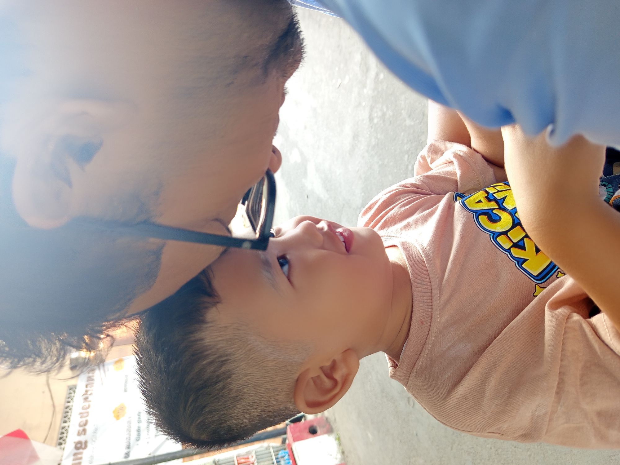 The media file is a close-up, low-angle shot of an adult and a child in what appears to be an outdoor setting. The adult, seen from the neck up, is wearing glasses and has dark hair. The adult's head is tilted down, facing the child. The child, whose face is prominently featured, has short hair and is looking up at the adult with a slight smile. The child is wearing a light pink t-shirt with a yellow and blue graphic design. The background is blurred, but some hints of a building or wall and a concrete ground are visible.
The interaction between the adult and child suggests a moment of connection, possibly a playful or affectionate exchange. The lighting indicates it is daytime, with bright sunlight casting some glare on the adult's face and hair. There are some faint, unreadable text elements on a sign in the background, to the left of the child. The overall scene conveys a sense of warmth and intimacy. The provided location context is Malang, Indonesia.