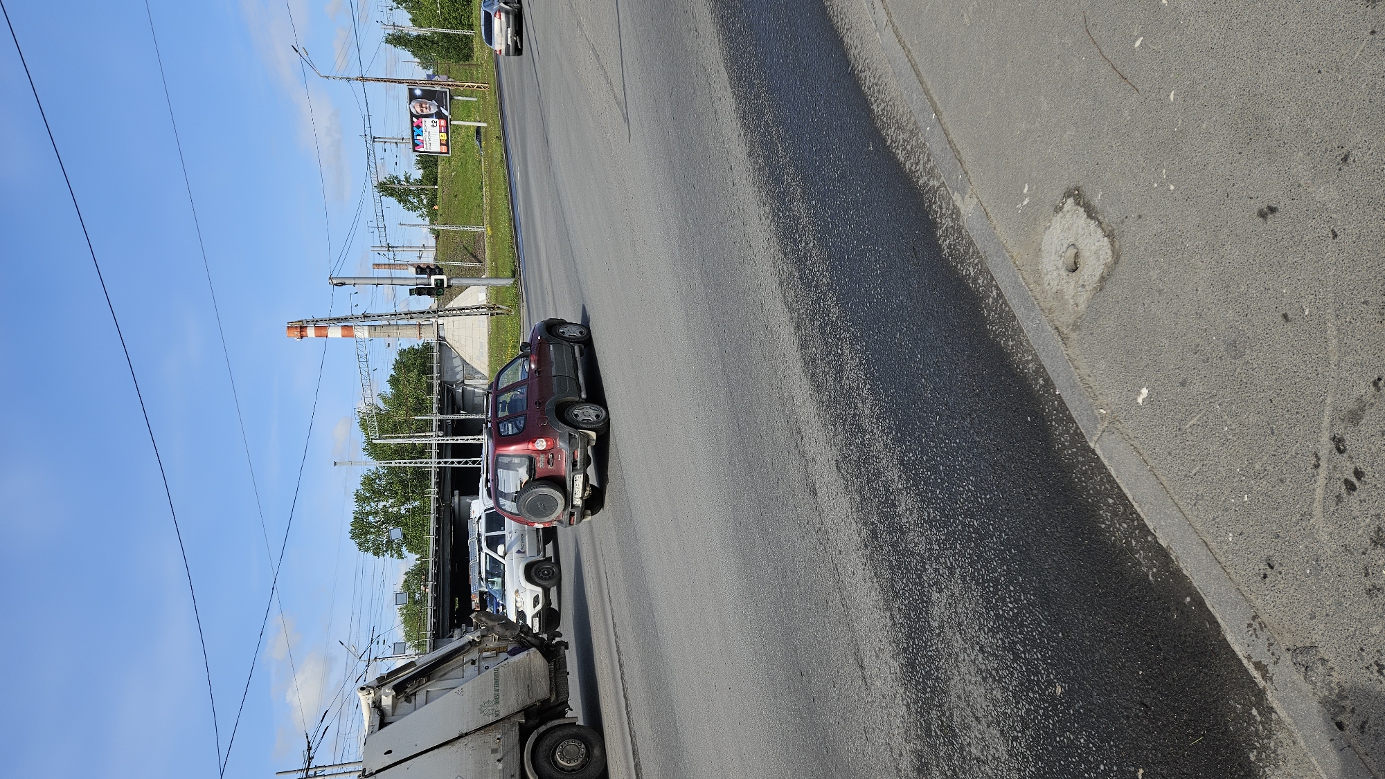 The image depicts a street scene with several vehicles parked along the roadside. The vehicles include a red SUV with a spare tire mounted on the rear, followed by a row of white cars. A large truck or utility vehicle is parked further down the street. In the background, there is a tall industrial chimney with red and white stripes, and overhead electrical wires are visible. A billboard is erected near a grassy area along the side of the road. The sky is blue with scattered clouds. The location appears to be an urban area, specifically in the city of Unknown, Unknown.