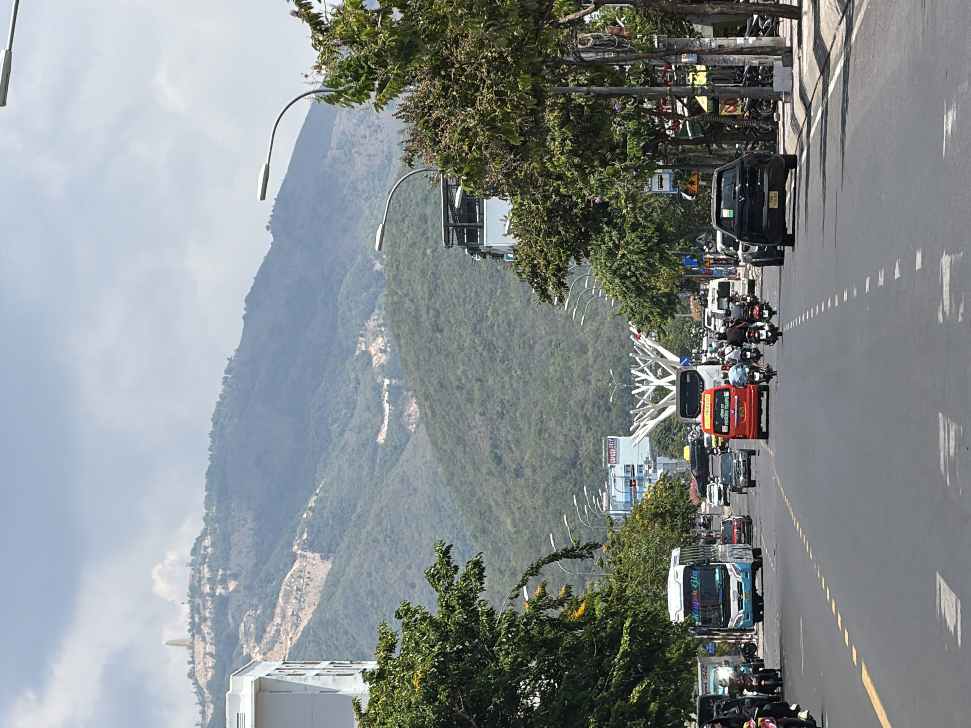 The image shows a street scene in Nam Nha Trang Ward, Vietnam, on a bright, possibly humid day. A multi-lane asphalt road with clear yellow and white lane markings dominates the foreground. Several vehicles are on the road, including a blue and white bus on the left with Vietnamese writing on its side, a red bus in the center, a black SUV in the right lane, and numerous motorcycles and smaller cars. On the sidewalks, there are trees with green foliage and some with yellow flowers, streetlights, and utility poles. Buildings are visible on the left and right sides of the road, including a light-colored building on the far left. In the background, lush green mountains rise, with a tall, slender pagoda-like structure visible atop one of the peaks. The sky is overcast with scattered clouds. The overall impression is a busy urban street scene with a natural mountainous backdrop.