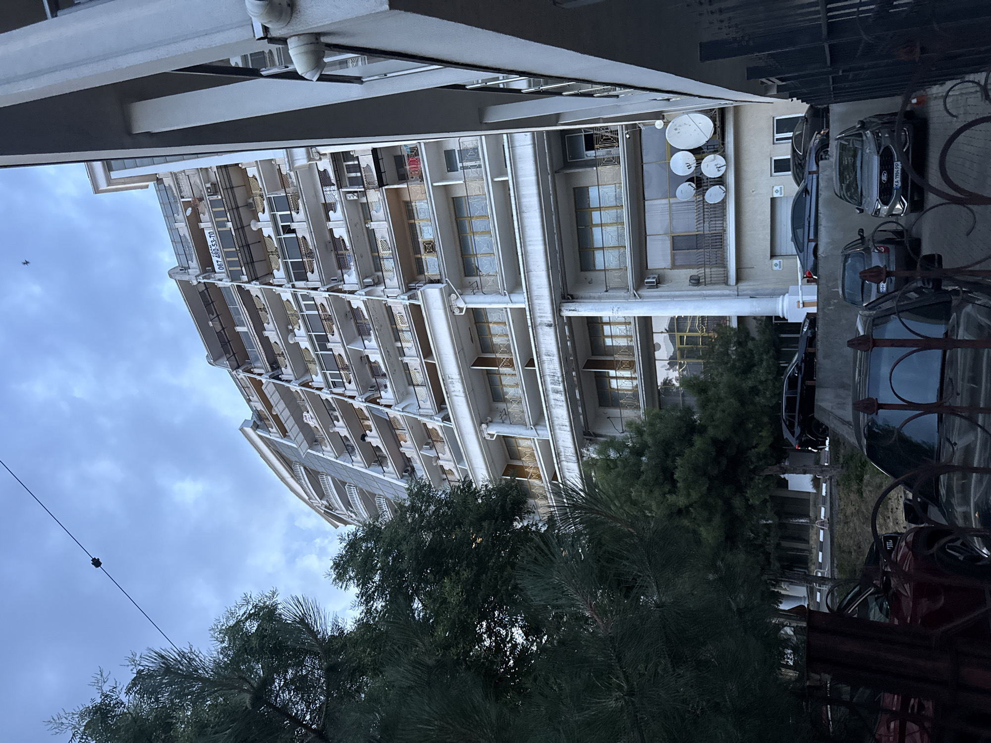 The image shows a multi-story apartment building in Odesa, Ukraine, with a distinctive curved facade and numerous balconies. A large billboard with the phone number "067 480-53-70" is prominently displayed on the building. The foreground features several parked cars, including a dark-colored SUV, a dark grey sedan, and a red SUV, suggesting a residential area. Evergreen trees, likely pine trees, are visible on the left side of the frame, partially obscuring the building. The sky is overcast with scattered clouds. The architectural style of the building appears to be modern, with a mix of white and light-colored materials, and golden accents on the balconies. Satellite dishes are mounted on some of the building's exterior walls. The overall impression is that of a contemporary urban environment.