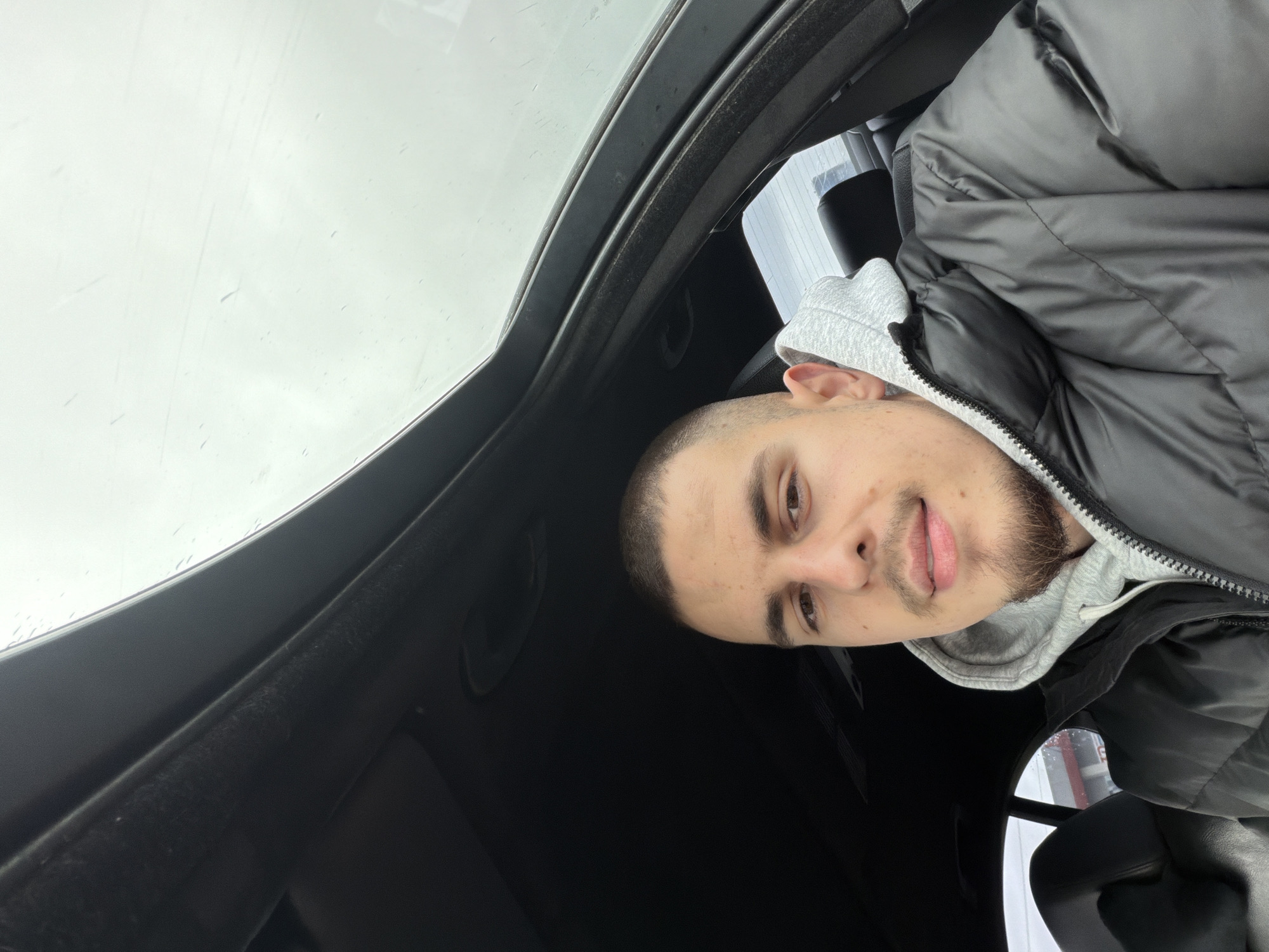 A man with short brown hair and a goatee is sitting in a car. He is wearing a grey hoodie and a black puffy jacket. The car has a sunroof. The sky is visible through the sunroof and appears to be overcast. It is impossible to determine the location in Plovdiv, Bulgaria from the image.