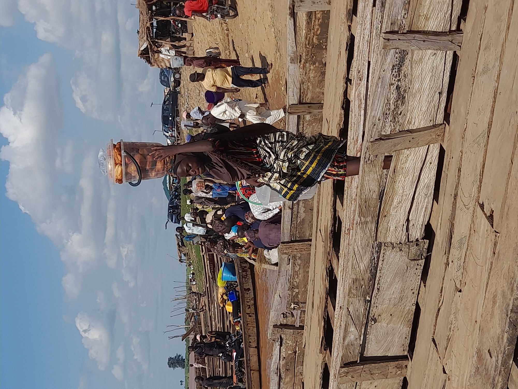 In the city of Ibi, Nigeria, a young woman is depicted from the chest up, carrying a large plastic bucket filled with food items balanced on her head. She is walking on a wooden platform, with a colorful, patterned skirt covering her lower body. The background reveals a bustling scene with multiple wooden boats docked along a riverbank. Several people are visible, some standing, others seated in or near the boats, suggesting a marketplace or a transportation hub. In the distance, more boats, structures, and vehicles can be observed under a bright, cloudy sky, indicating a vibrant, active environment.