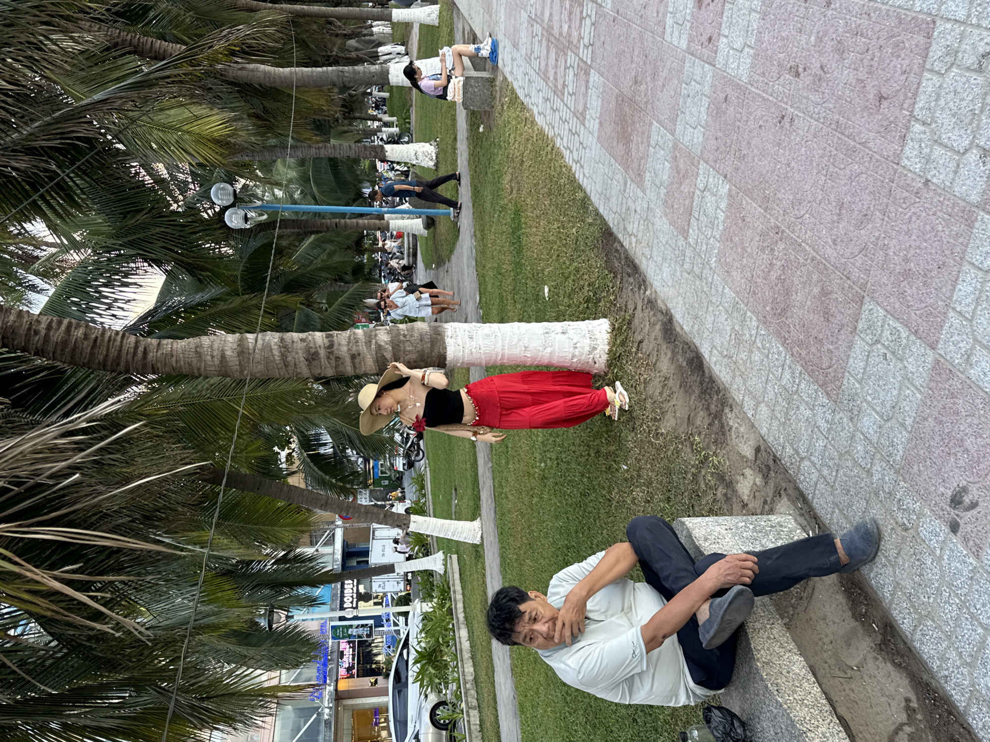 In Nha Trang Ward, Vietnam, an outdoor scene unfolds with a man seated on a concrete bench in the foreground, his hand near his mouth. Behind him, a woman in a black top and a long red skirt poses next to a palm tree, her hand holding a straw hat. The environment features a grassy area with more palm trees, a paved walkway with a distinctive checkered pattern of light gray and pink tiles, and buildings in the background. Several people are visible in the midground and background, walking and sitting, along with parked motorcycles and cars. The lighting suggests it is late afternoon or early evening.