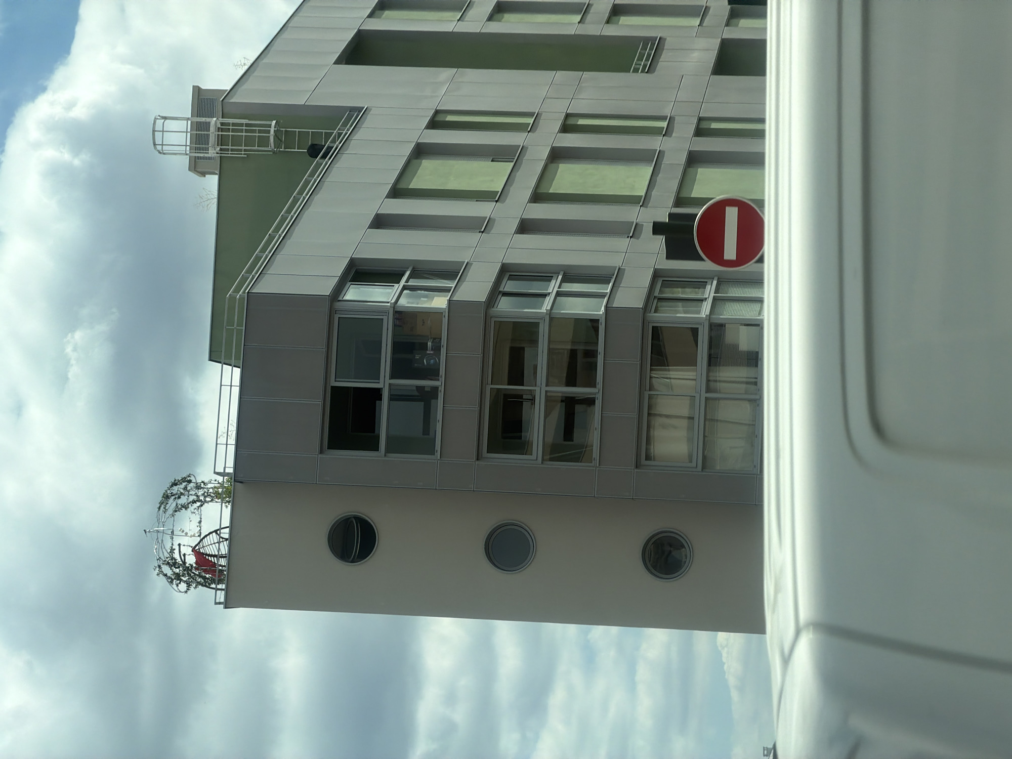The media file shows a modern building with a gray facade and green-tinted windows. The building has several floors, with a balcony on the lower level featuring a decorative, crown-like structure adorned with vines and a red chair. Three circular windows are visible on the lower section of the building. A red and white "no entry" sign is mounted on the building's facade. The sky in the background is cloudy. The image appears to have been taken during the daytime. A portion of a white vehicle is visible in the foreground, partially obscuring the view of the building. The image is oriented vertically, with the building appearing to lean to the left. Based on the provided location context, this scene is in Alfortville, France.