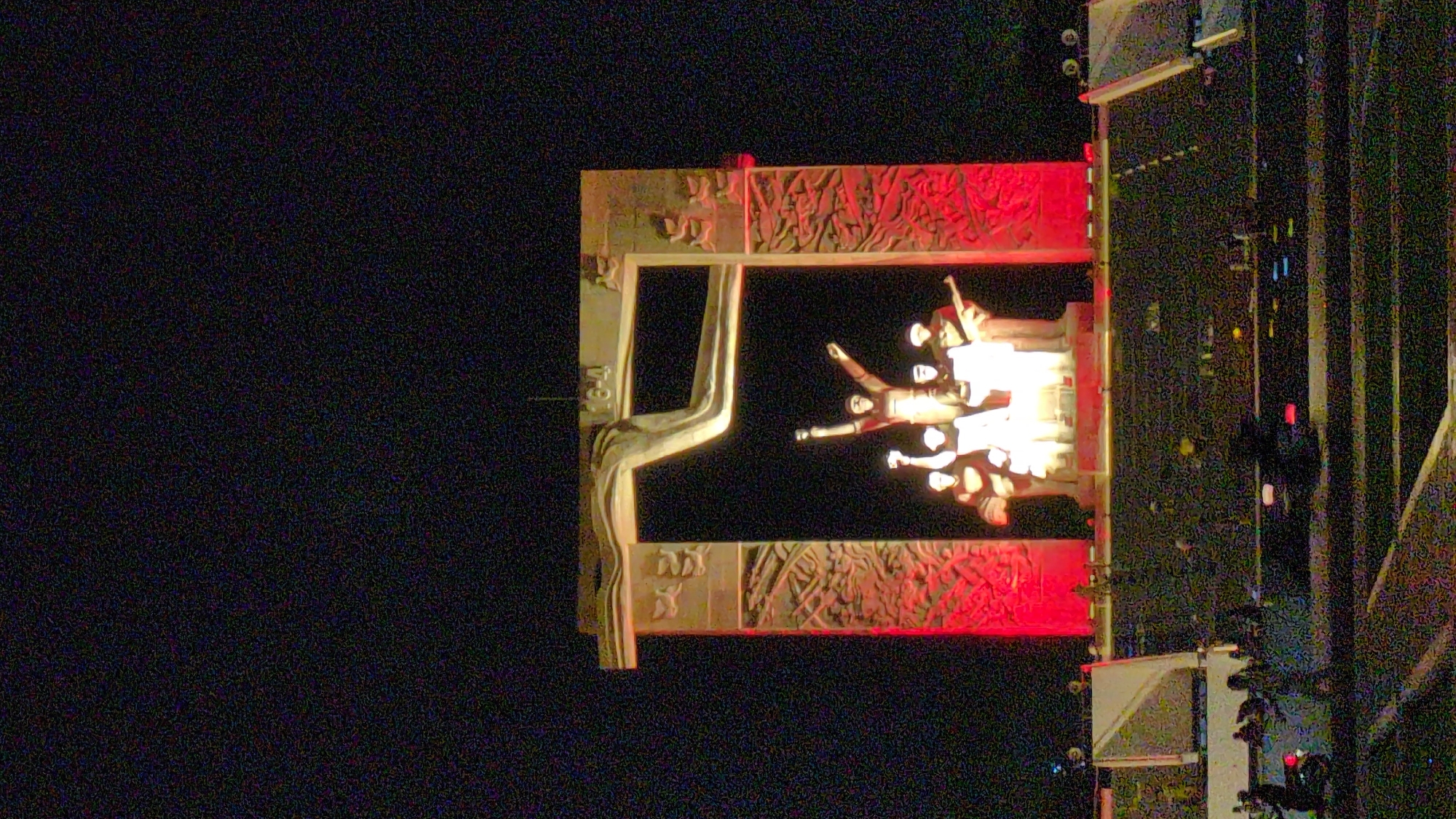 At night in Phan Rang – Tháp Chàm, Vietnam, a sculpture stands illuminated. It features a large frame decorated with carvings. Within the frame, a group of figures appear, brightly lit. A large, stylized arm extends from the left side of the frame. A road and structures are visible to the right of the sculpture. The background is dark.